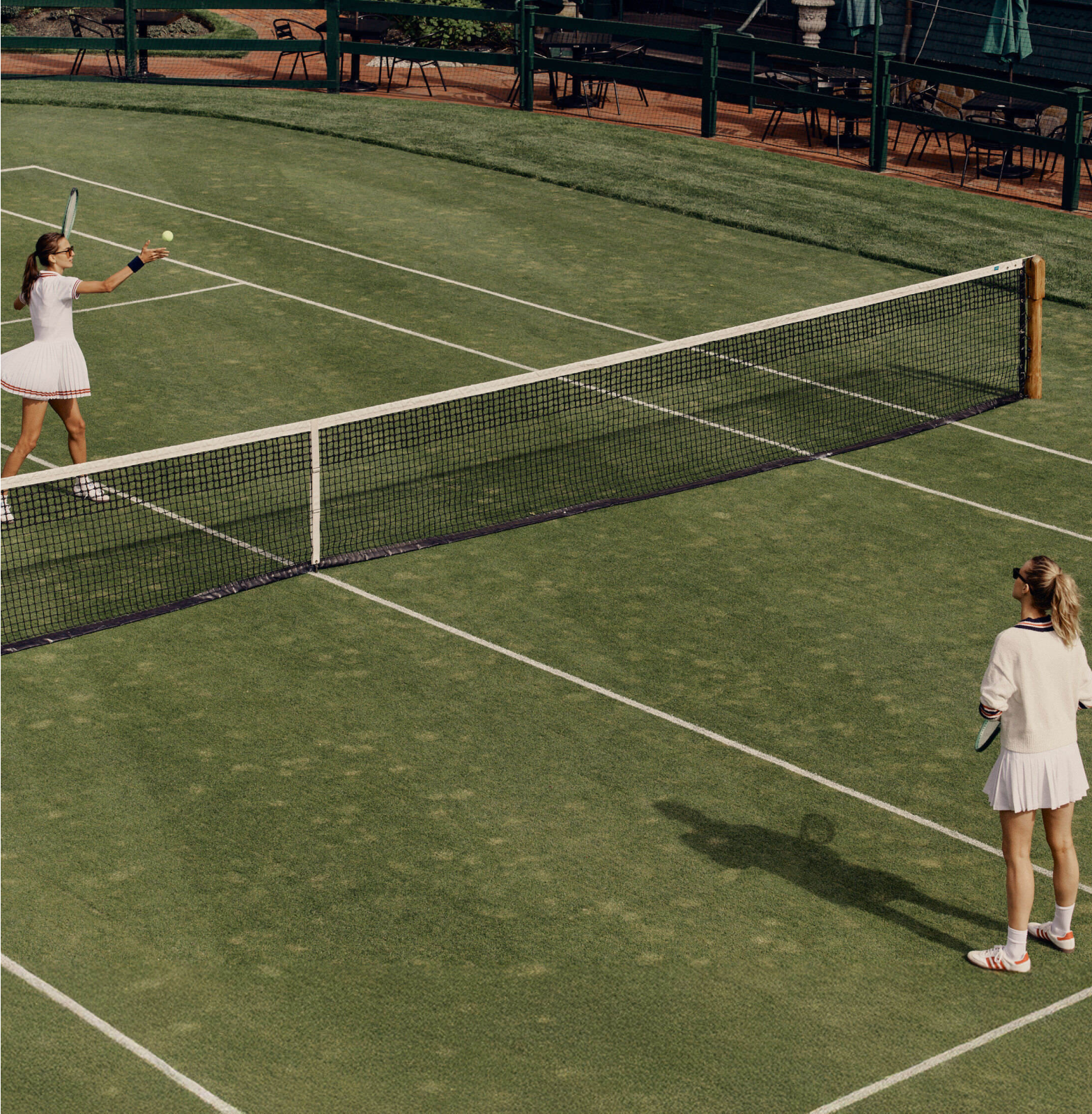 Two female tennis players in classic white attire engage in an effortless, high-end grass-court match, embodying fun and confident style.