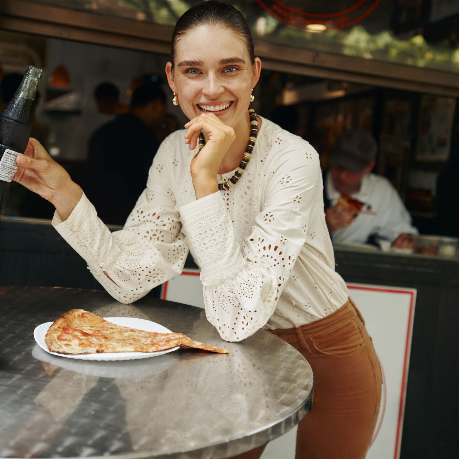 A young person in a classic, embroidered blouse enjoys an effortless, fun moment with pizza and a soda, embodying Metrobazaarstore's high-end, relaxed style.