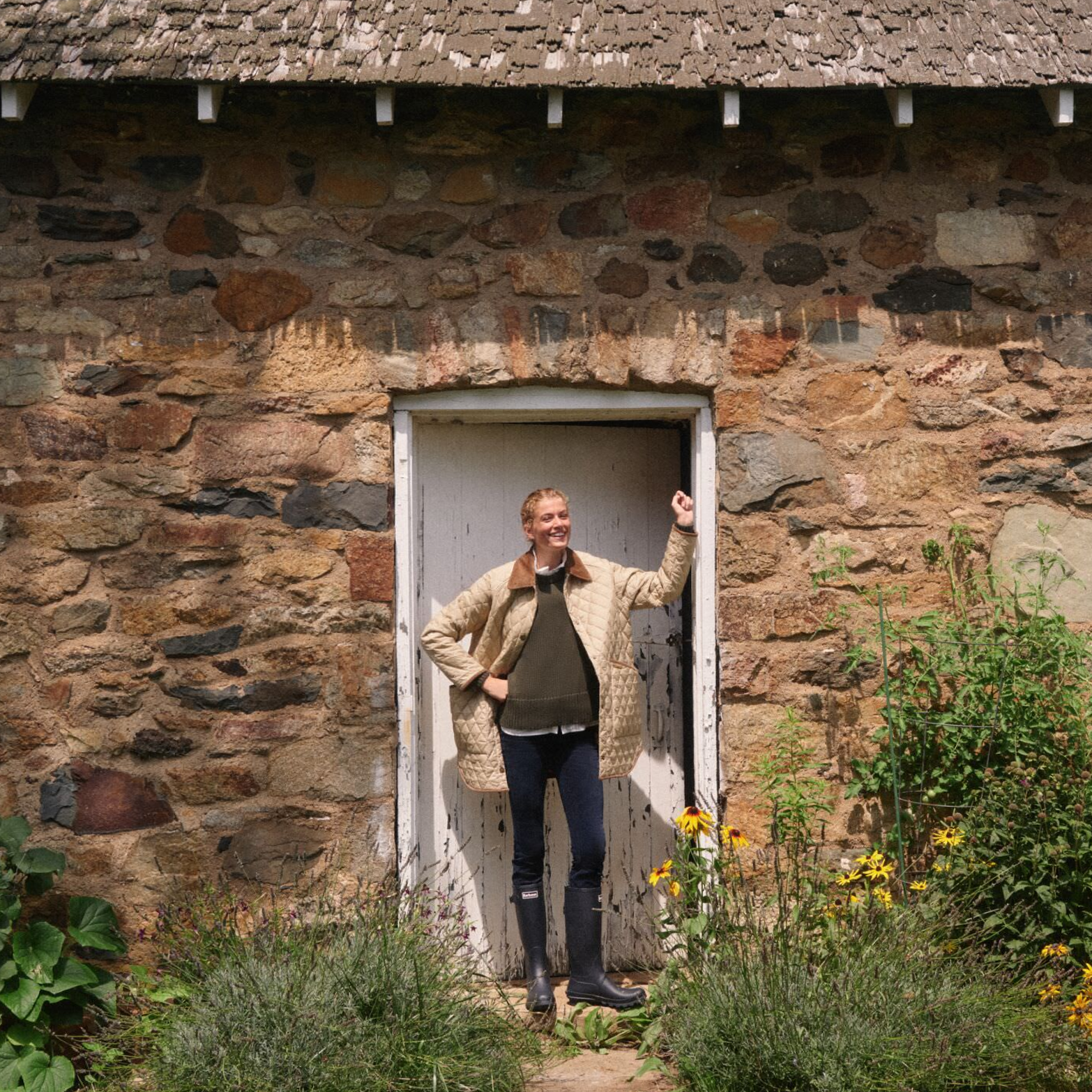 Person in a classic, quilted beige coat, stands in rustic doorway surrounded by flowers, embodying Metrobazaarstore's effortless, fun, and high-end style.