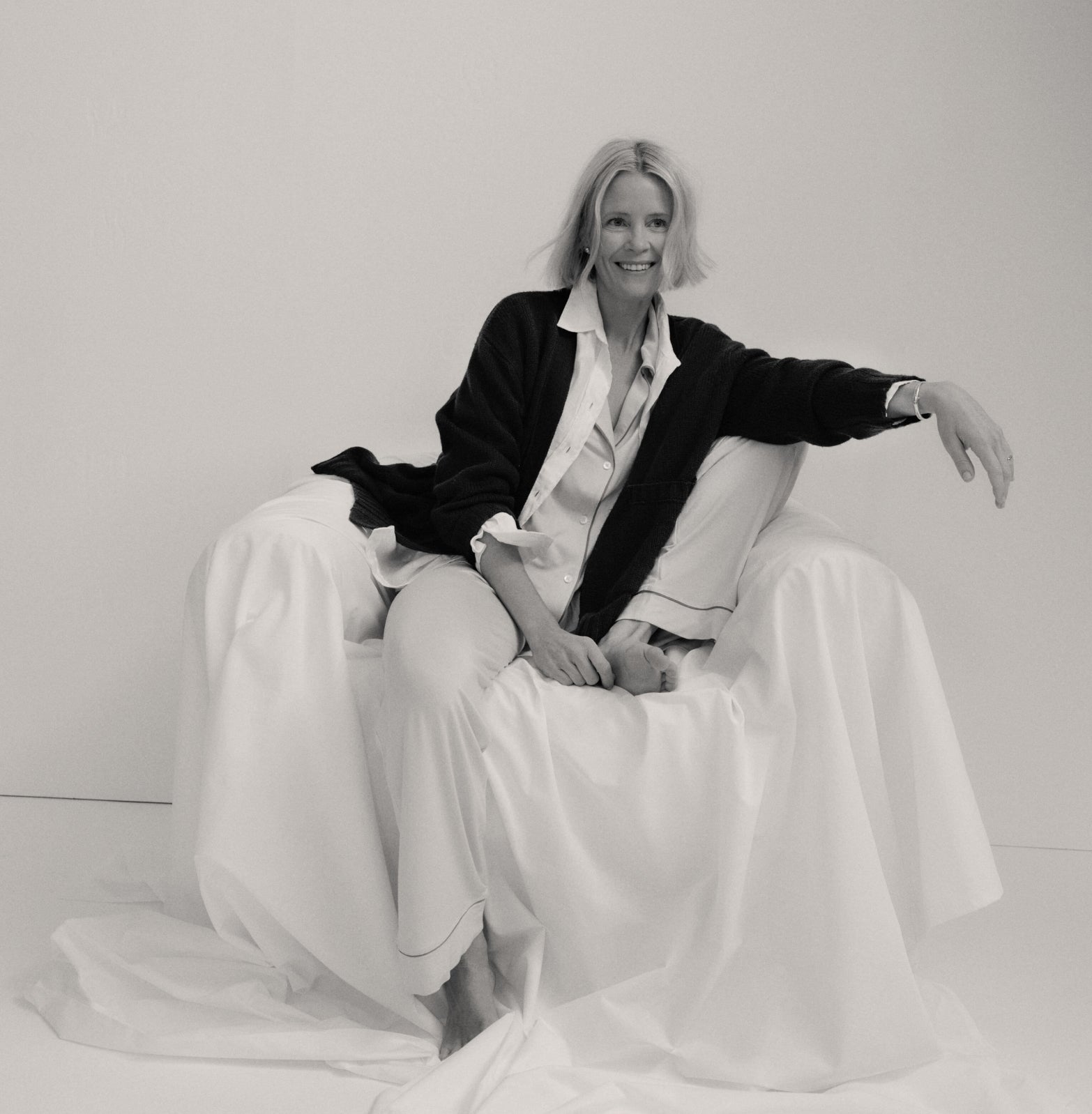 Black-and-white photo of a woman in classic, loose-fitting pajamas and cardigan, seated effortlessly on a draped armchair, exuding a high-end, relaxed vibe.