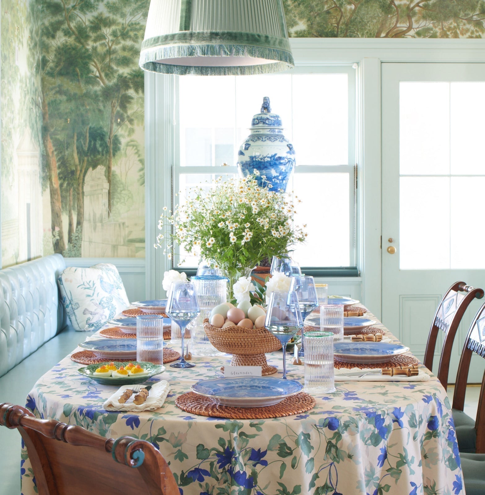 Spring-inspired dining table with cream floral tablecloth, blue-and-white plates, pastel eggs, daisies, and hors d’oeuvres, exuding classic, effortless, high-end fun.
