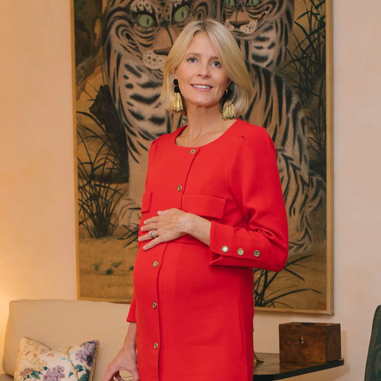 A woman in a classic red button-front coat, effortlessly accessorized, stands indoors, highlighting her pregnant belly. High-end decor and art enhance the scene.