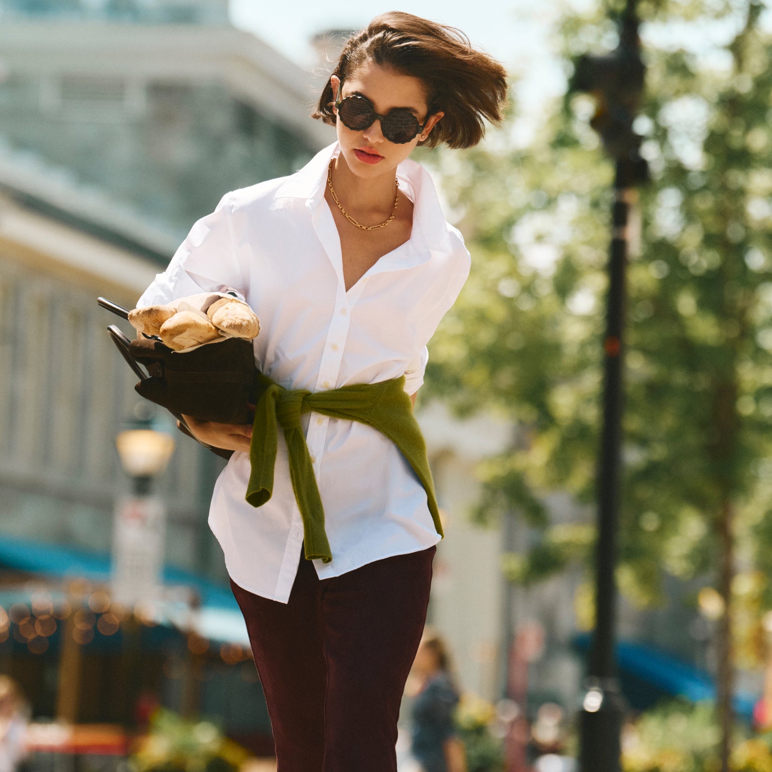 A casually chic woman embodies effortless style in a classic white shirt, light green sweater, and dark pants, carrying a tote with fresh baguettes.