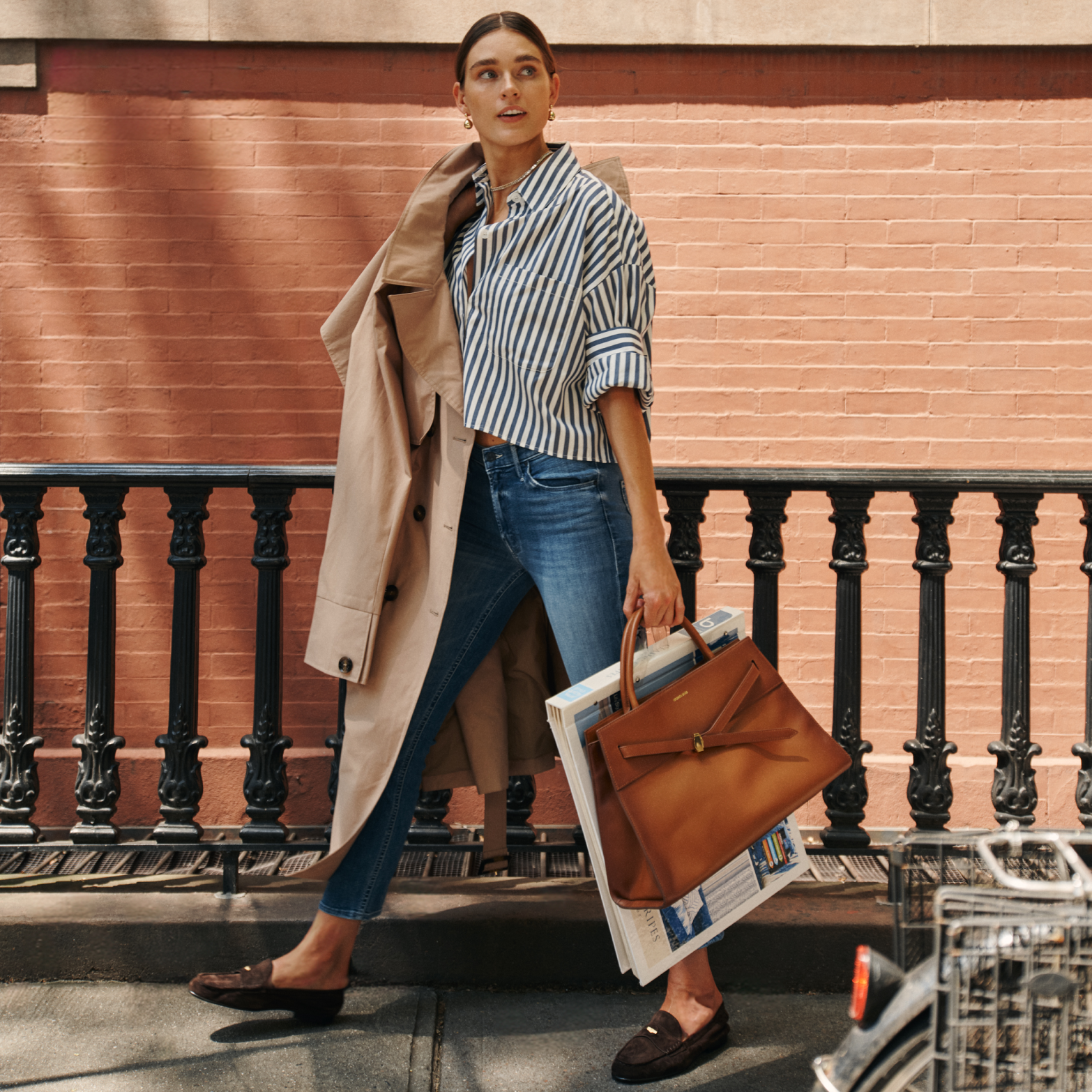 Woman in classic, effortless style with a trench coat, blue jeans, and loafers, carrying a high-end leather tote, exuding fun confidence.