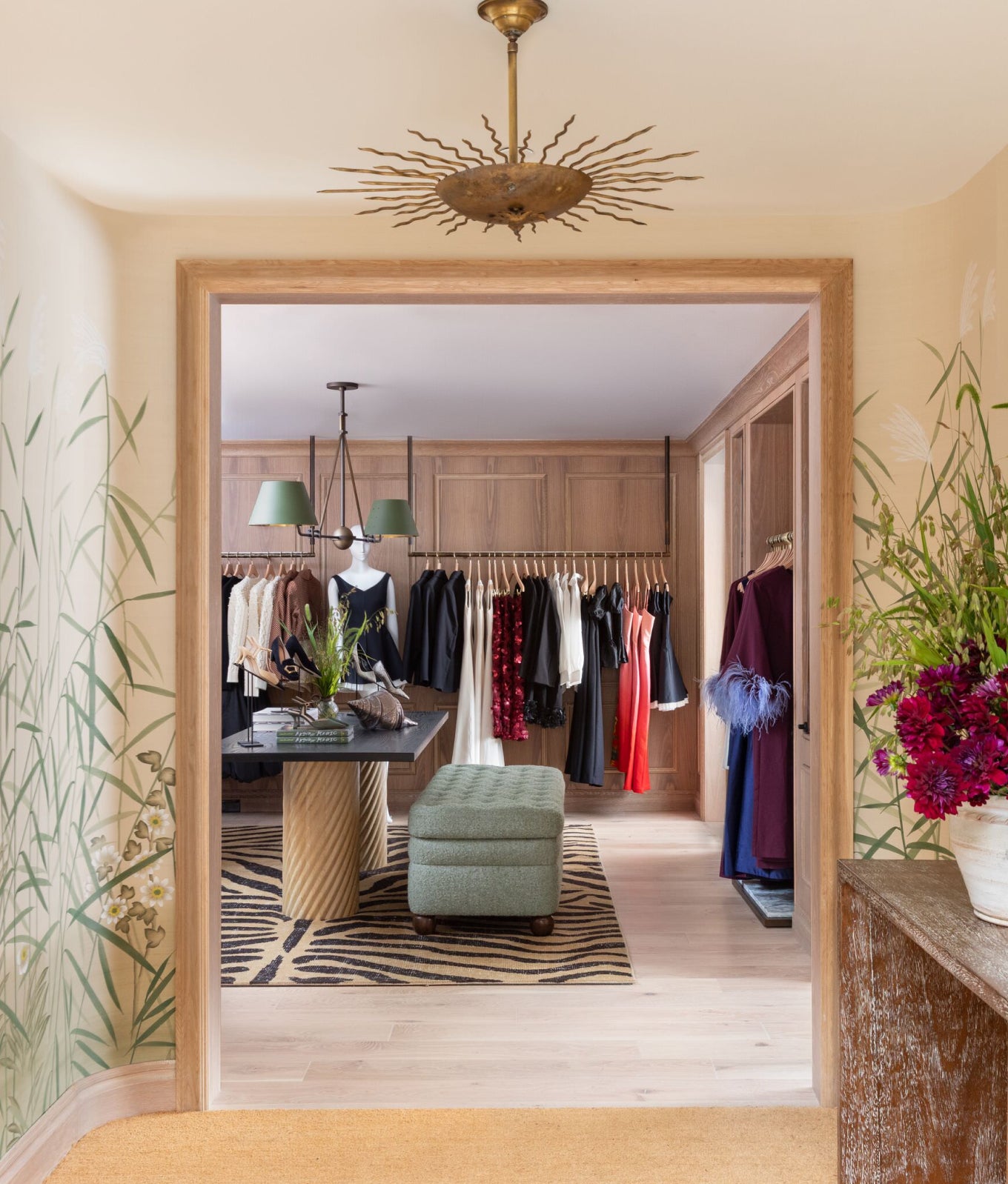 A classic, high-end dressing room with effortless style: wooden cabinets, garment racks with colorful clothes, green ottoman, zebra rug, chandelier, and a console with flowers.