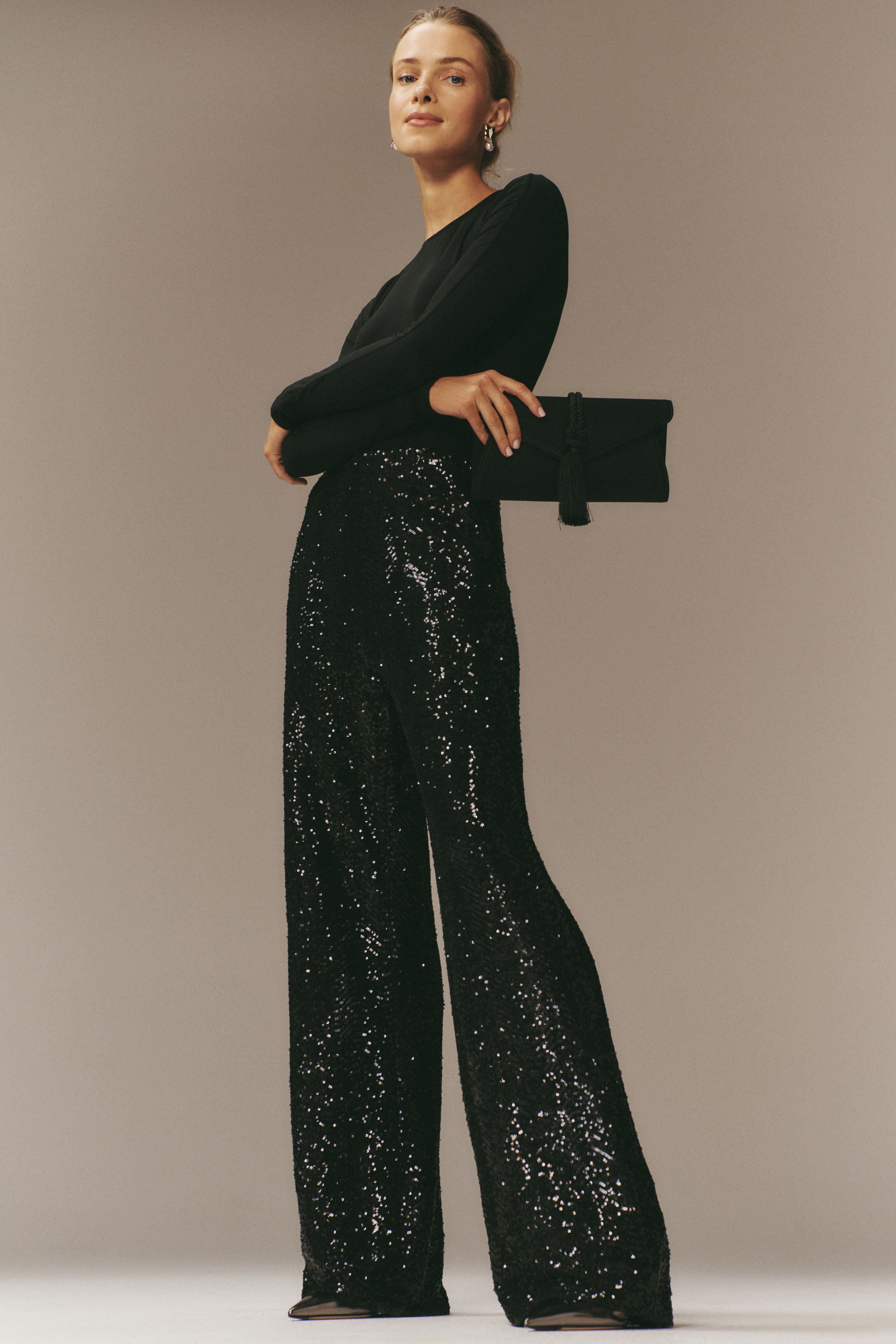 Model wearing classic Black Sequin Tatum Pants with a high-rise, wide-leg silhouette, paired with a fitted black top and clutch, exuding effortless, high-end evening elegance.