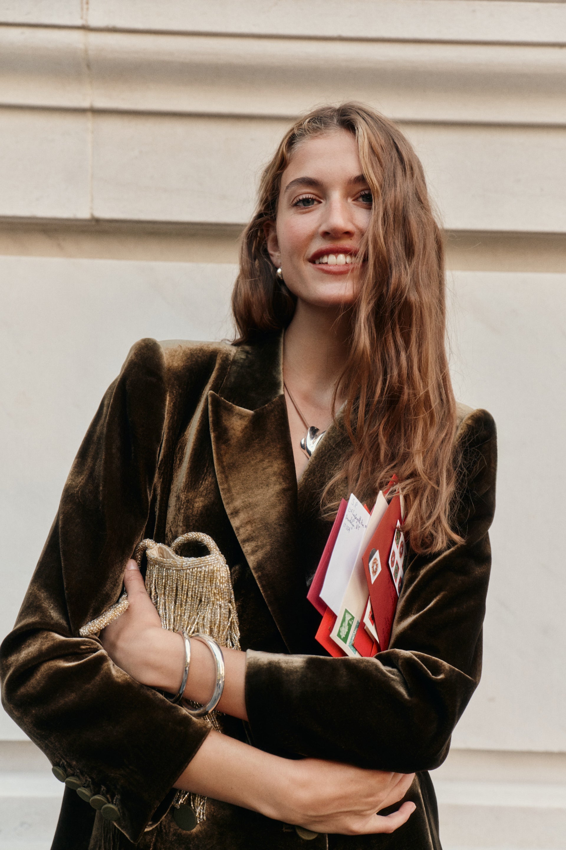 A woman wearing a dark olive velvet blazer holds a gold fringe purse and a collection of mail pieces.