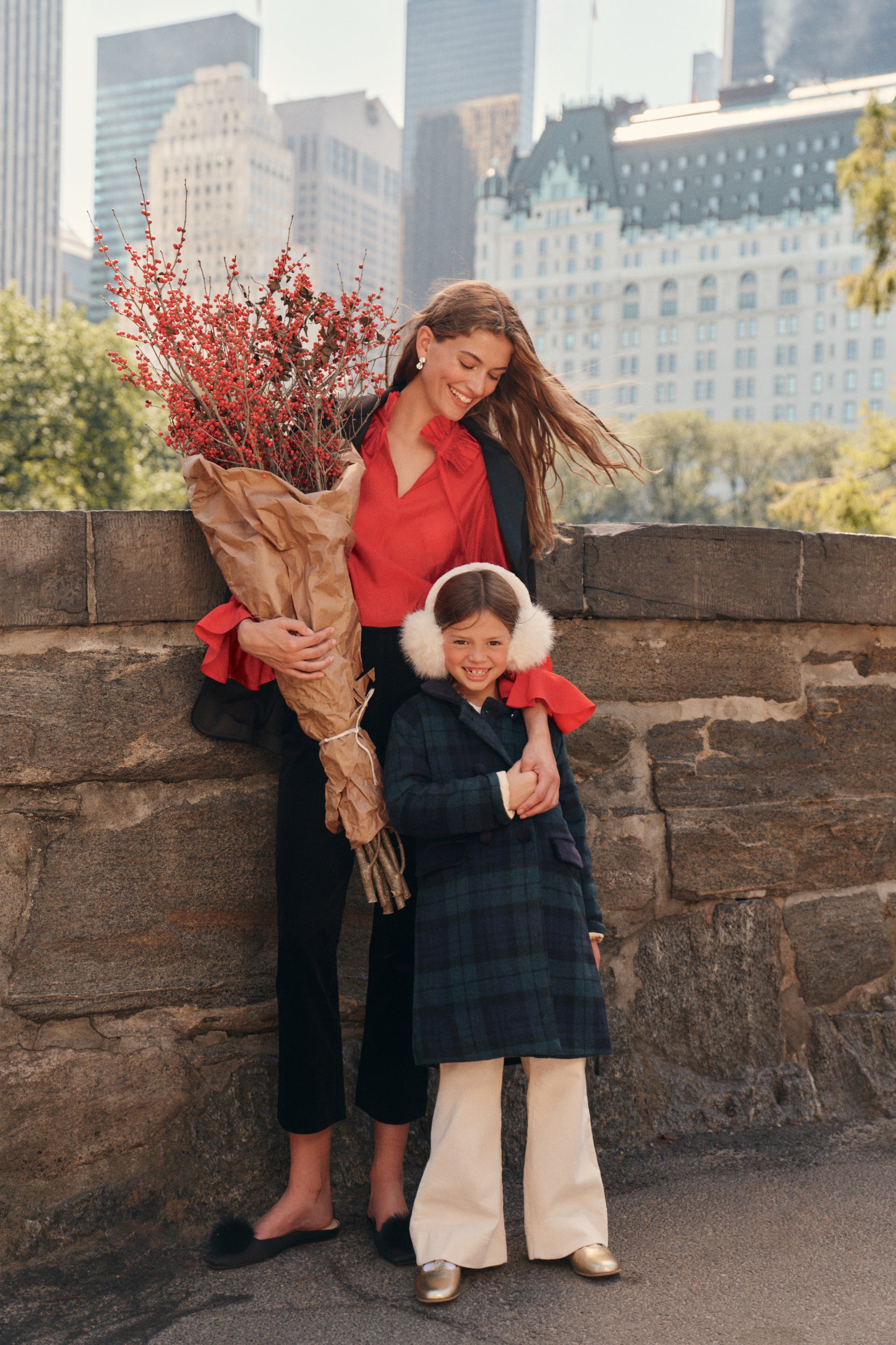 A woman stands on a city park bridge with a little girl. The woman wears a black blazer, a red blouse, black pants, and black flats while carrying a set of paper wrapped florals. The little girl wears a tartan patterned coat, cream pants, gold t-strap shoes, and ear muffs.