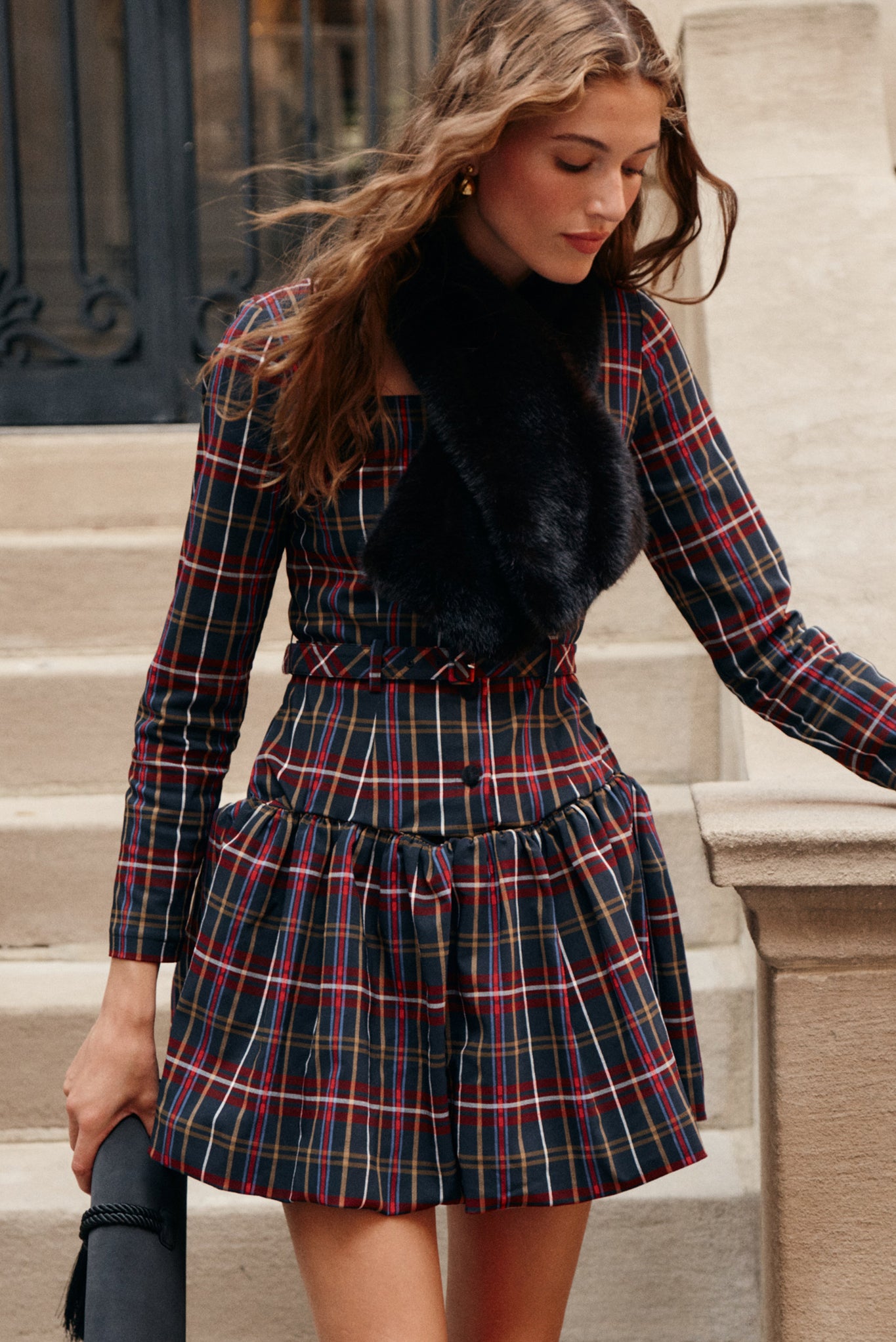 A woman wearing a tartan patterned mini dress and a brown fur scarf carries a black tassel clutch.
