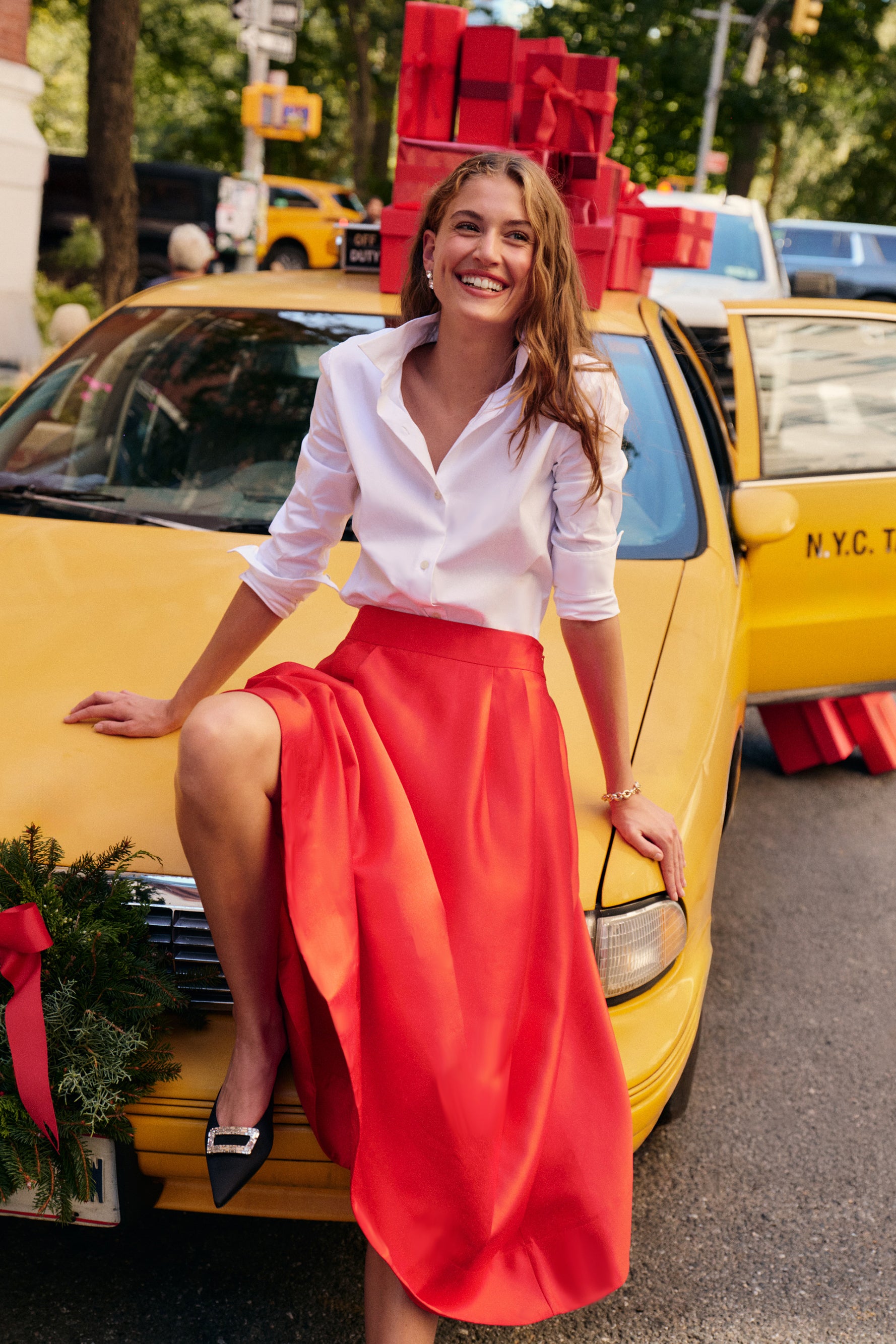 A woman leans against a yellow city cab wearing a white button down, a red dupioni midi skirt, and black heels.