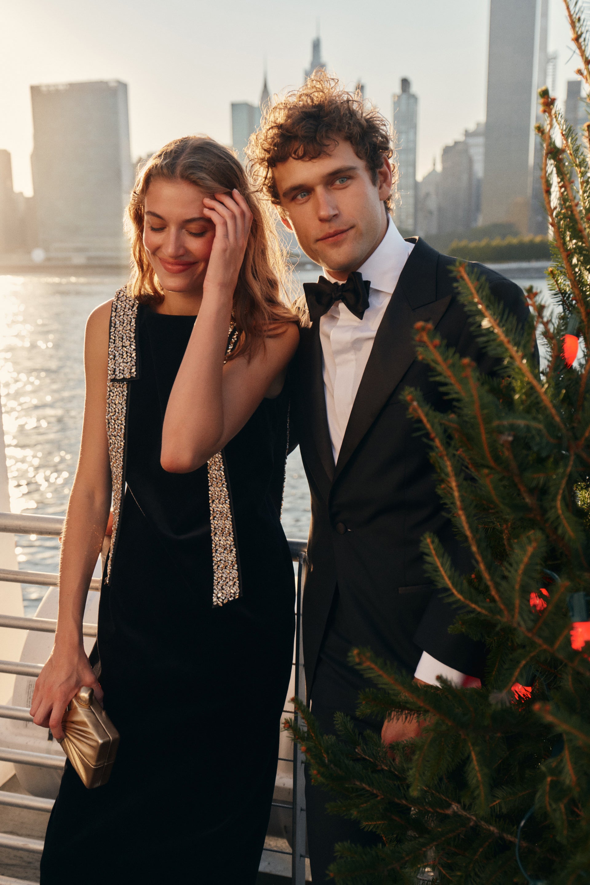 On the left, a woman wears a black midi dress with embellished shoulder bows and carries a gold clutch. On the right, a man wears a suit and bow tie next to a Christmas tree.