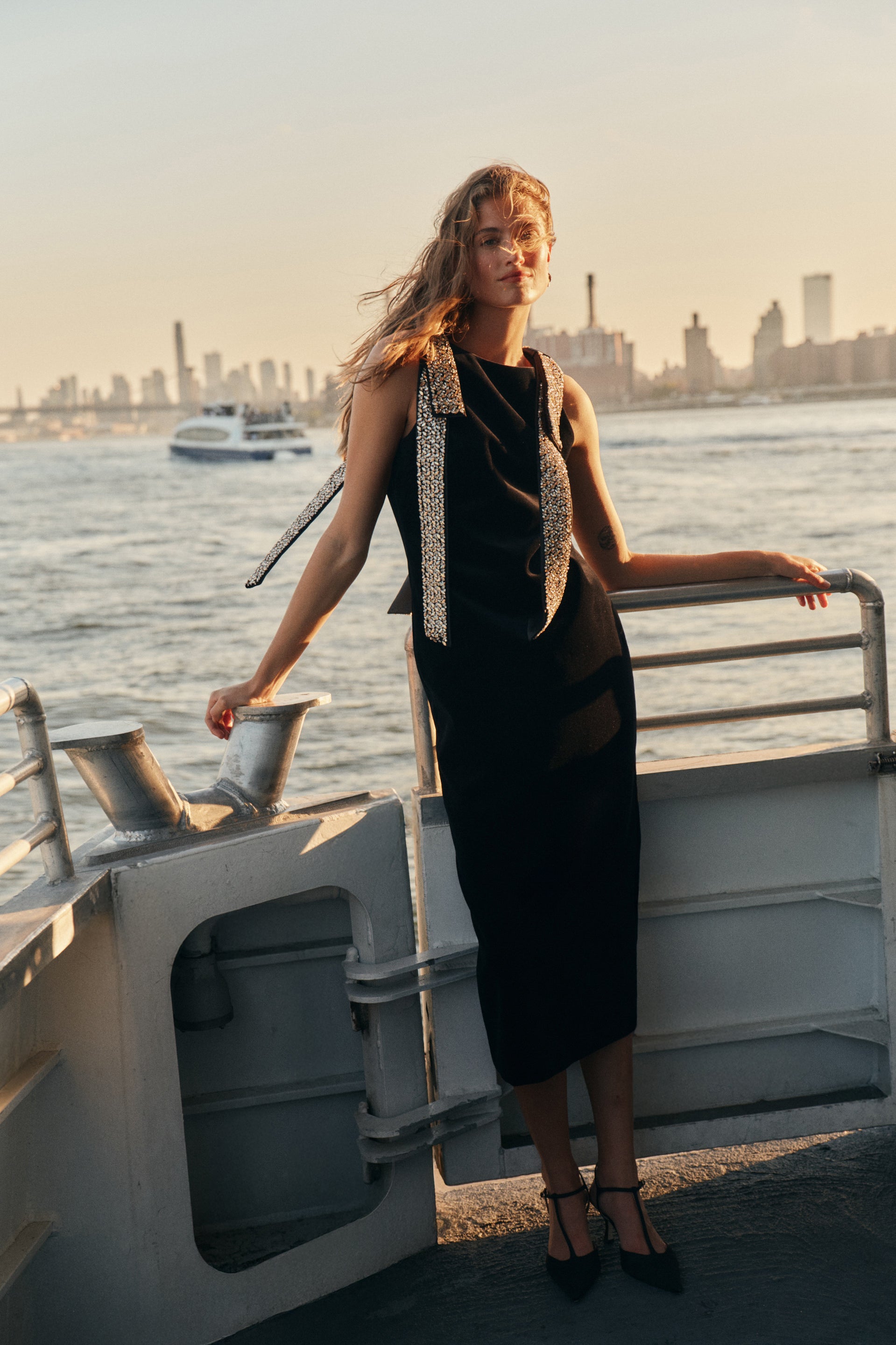 A woman on a ferry wearing a black midi dress with embellished shoulder bows and black t-strap heels.