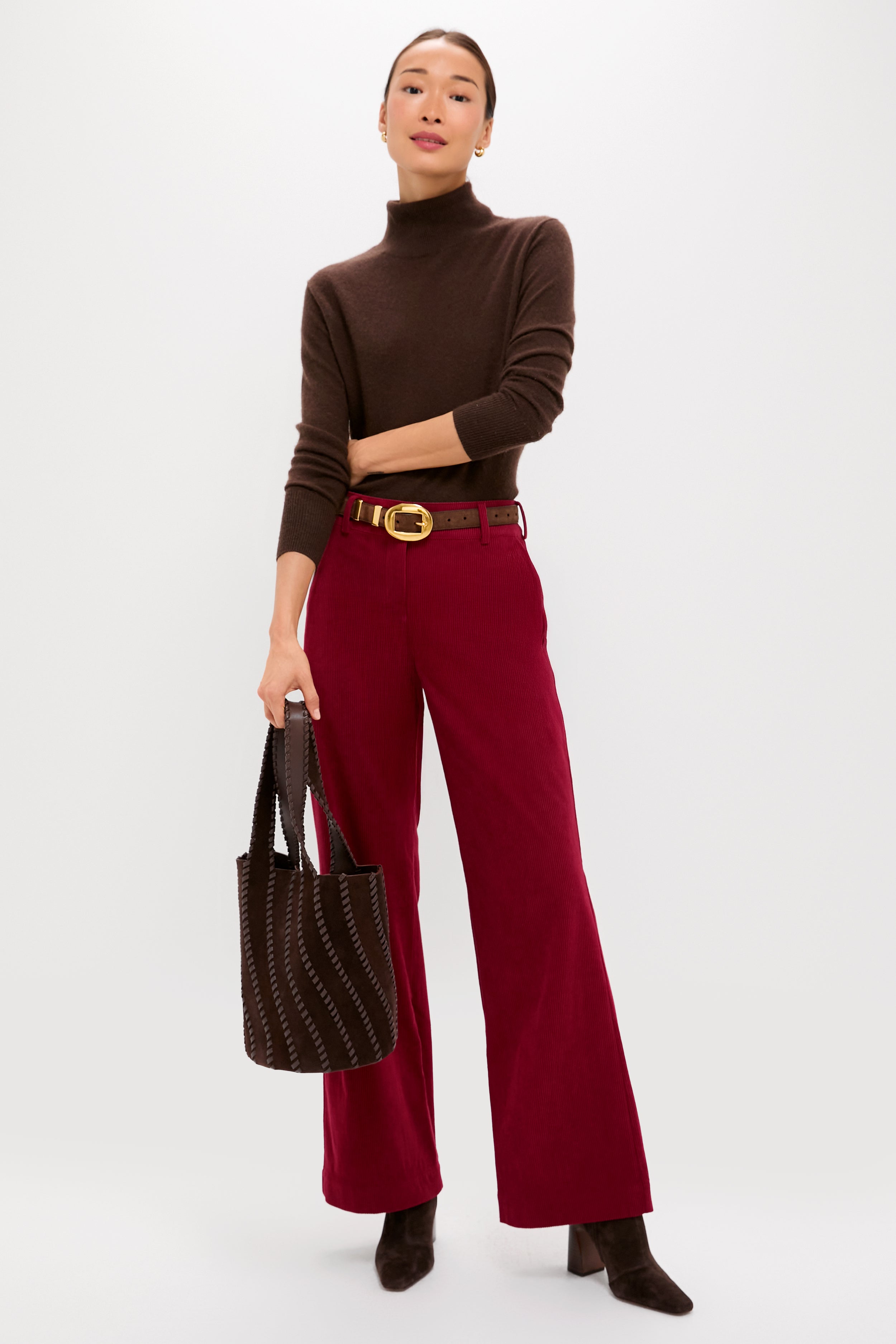 A woman wears a Metrobazaarstore brown turtleneck, wine wide-leg pants, brown boots, a gold-buckled belt, and holds a brown tote bag against a plain background.