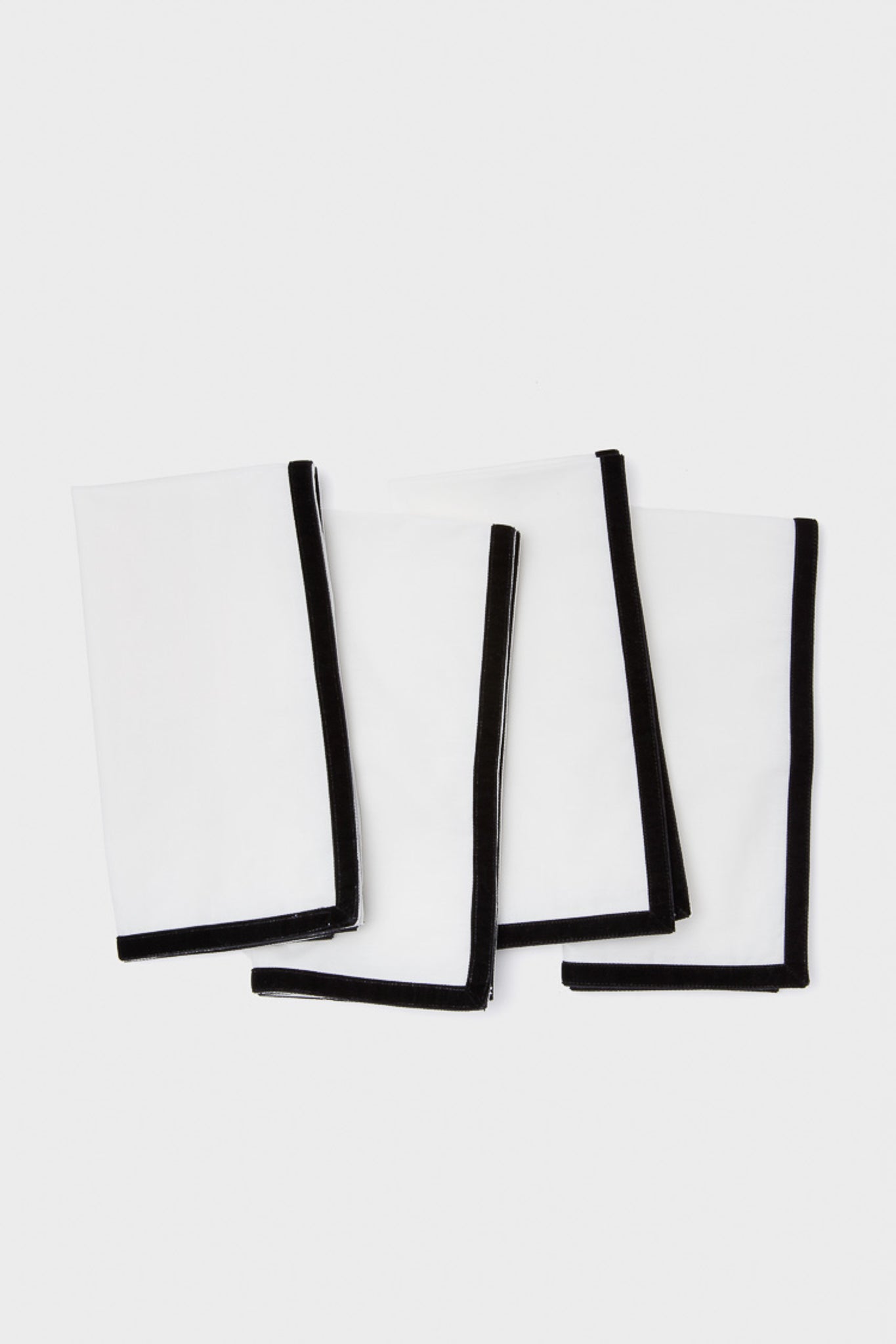 Four white cotton napkins from Metrobazaarstore Home, featuring elegant black velvet trim, are neatly arranged in an overlapping pattern on a plain white background.