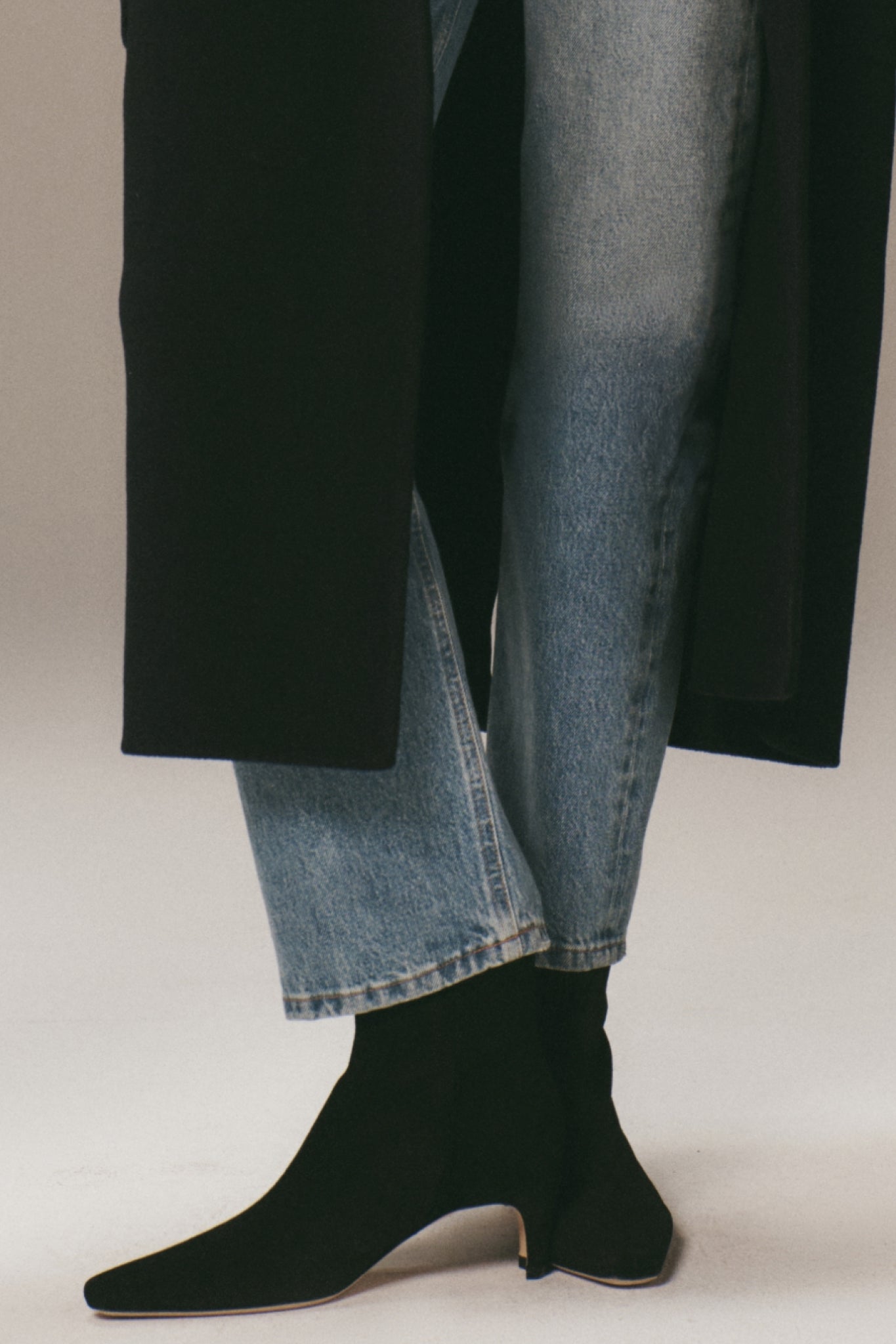 Model wearing classic Black Suede Wally Ankle Boots with pointed square toe and low heel, paired effortlessly with cropped jeans and a black overcoat. High-end, fun style.