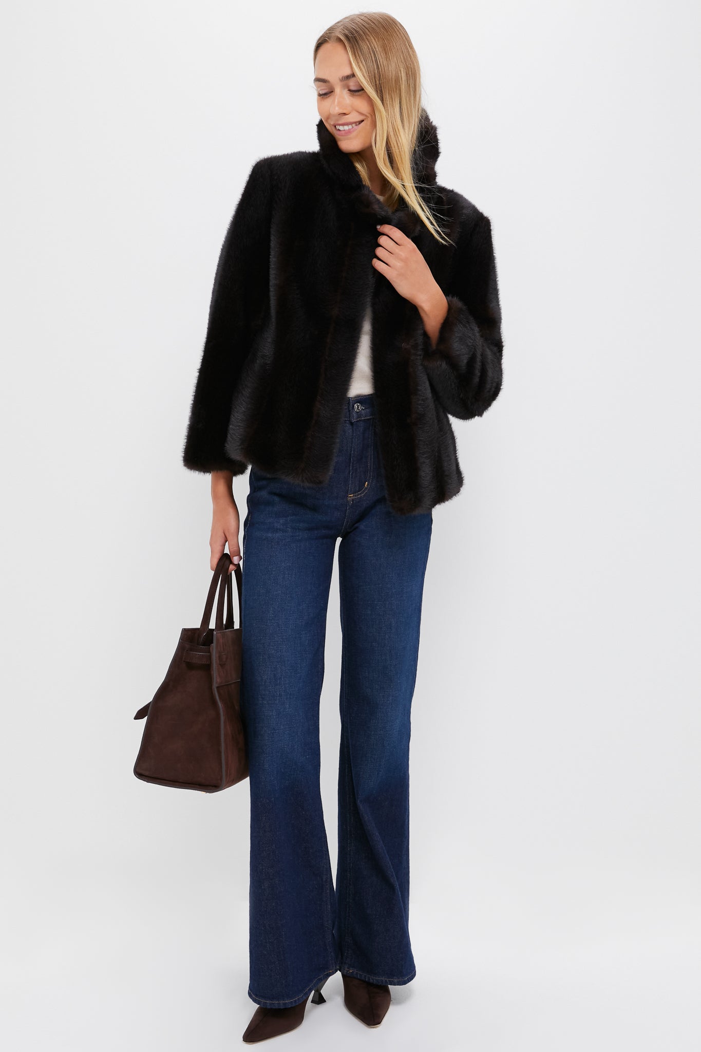 A woman in a Metrobazaarstore black faux fur coat, white top, blue wide-leg jeans, and brown heels holds a dark brown handbag against a plain white background—an effortlessly chic winter statement look.