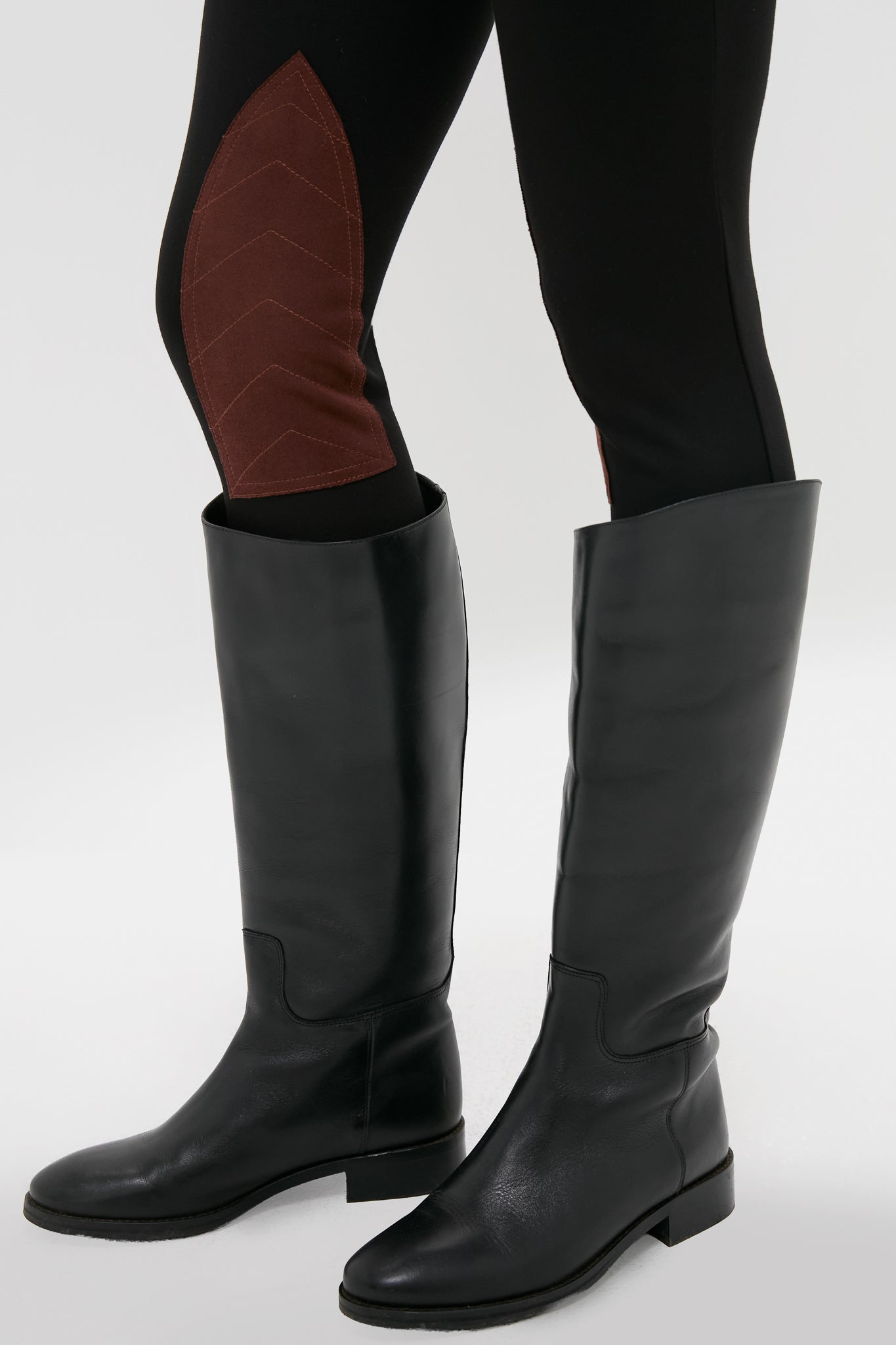 A person wearing Metrobazaarstore Shoes black knee-high leather boots and black leggings with brown chevron-patterned patches on the inner knees.
