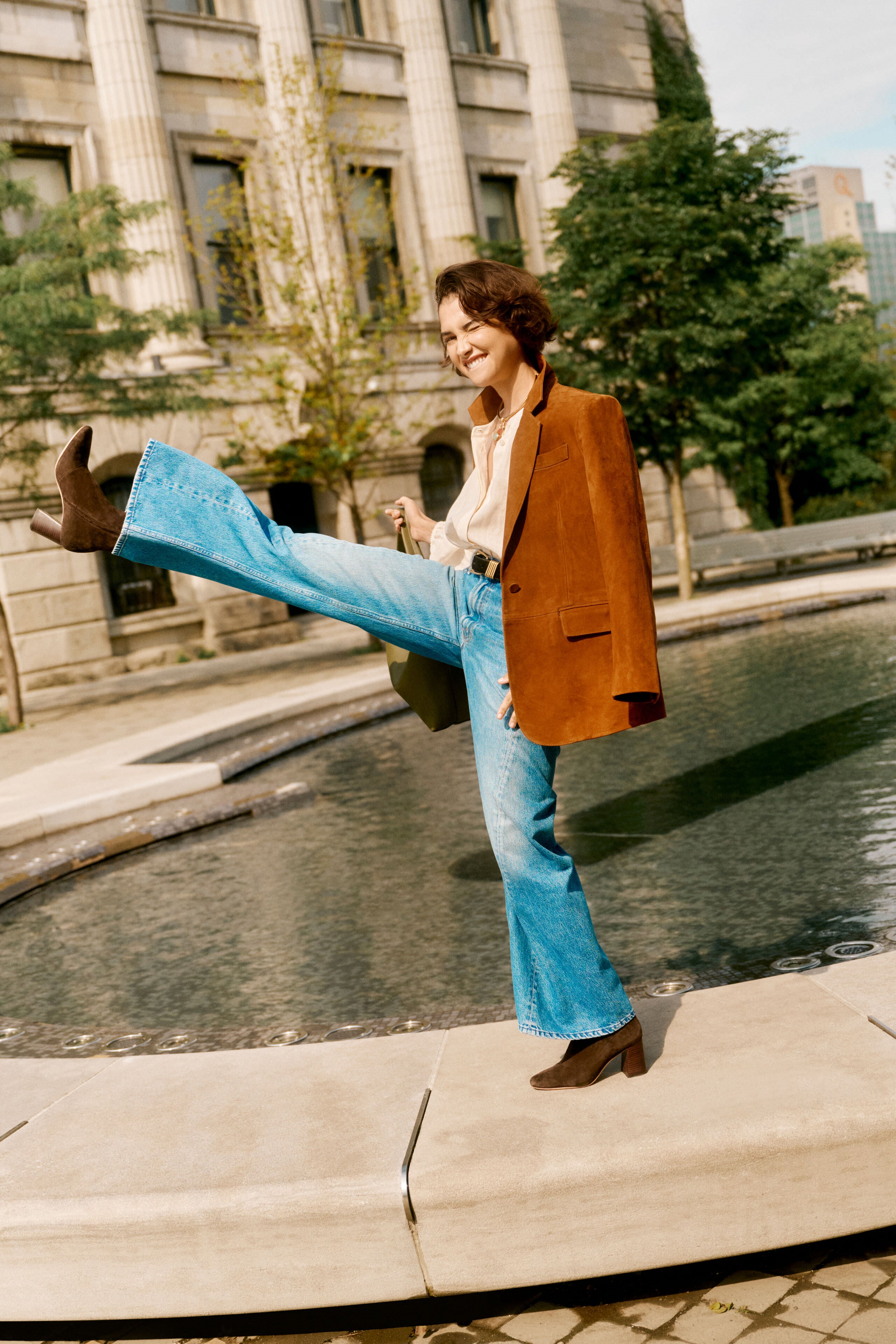 Young person playfully poses by a fountain, wearing classic Tmoro Stretch Suede Chloe Boots, effortlessly paired with high-waisted jeans and a blazer in a fun, urban setting.