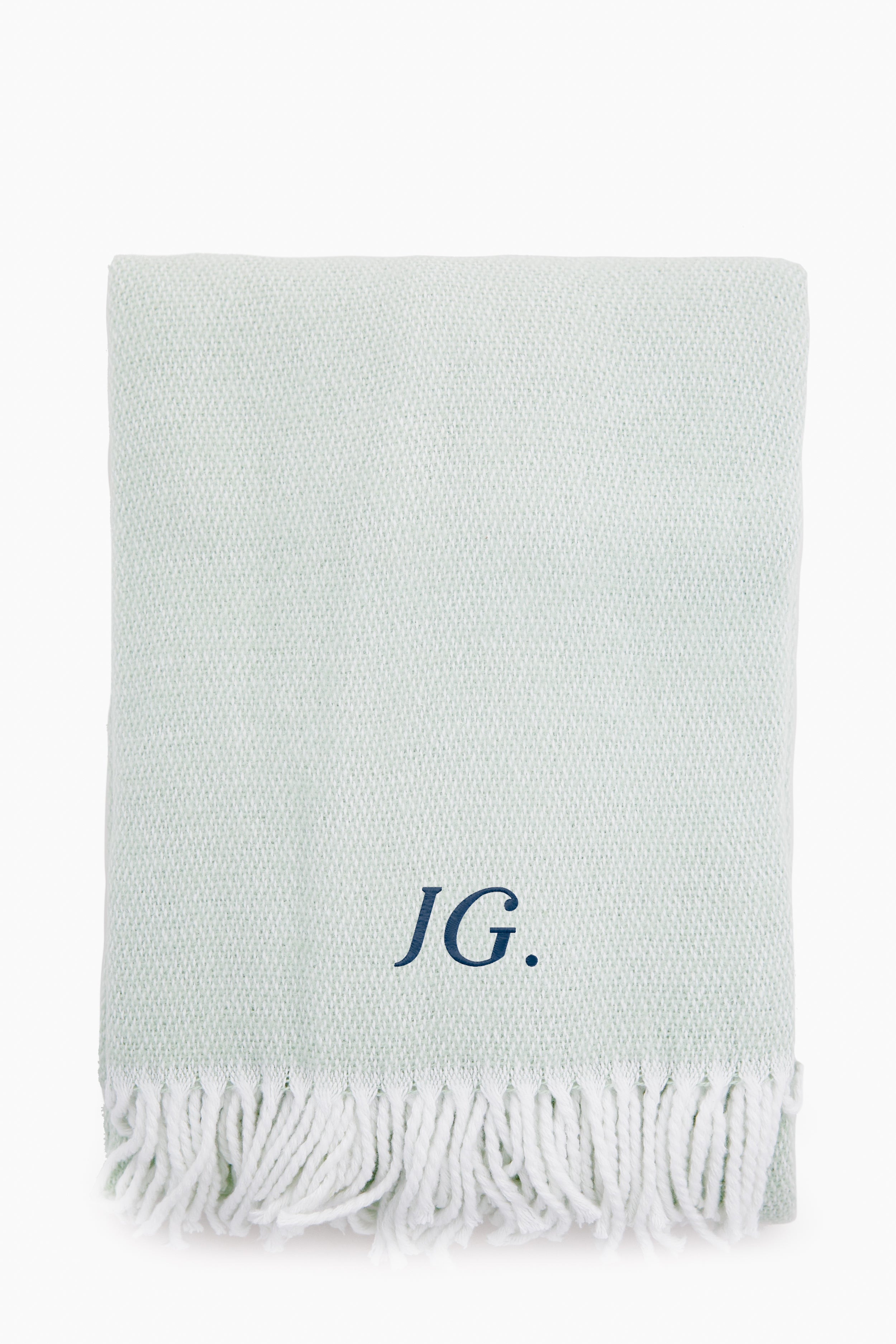 A light gray Matouk Opal Pezzo Throw features a white fringe and the initials JG. embroidered in dark blue on the front. Made from soft cotton, this elegant blanket adds style to any space.