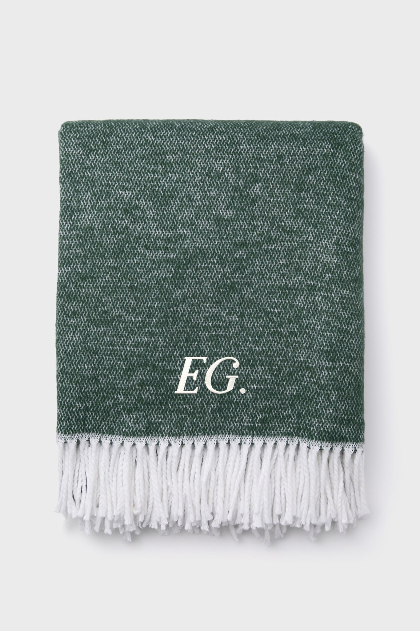A folded green Jade Pezzo Throw by Matouk, featuring a textured finish, white fringed edges, and the initials E.G. in white on one side, adds an elegant touch.