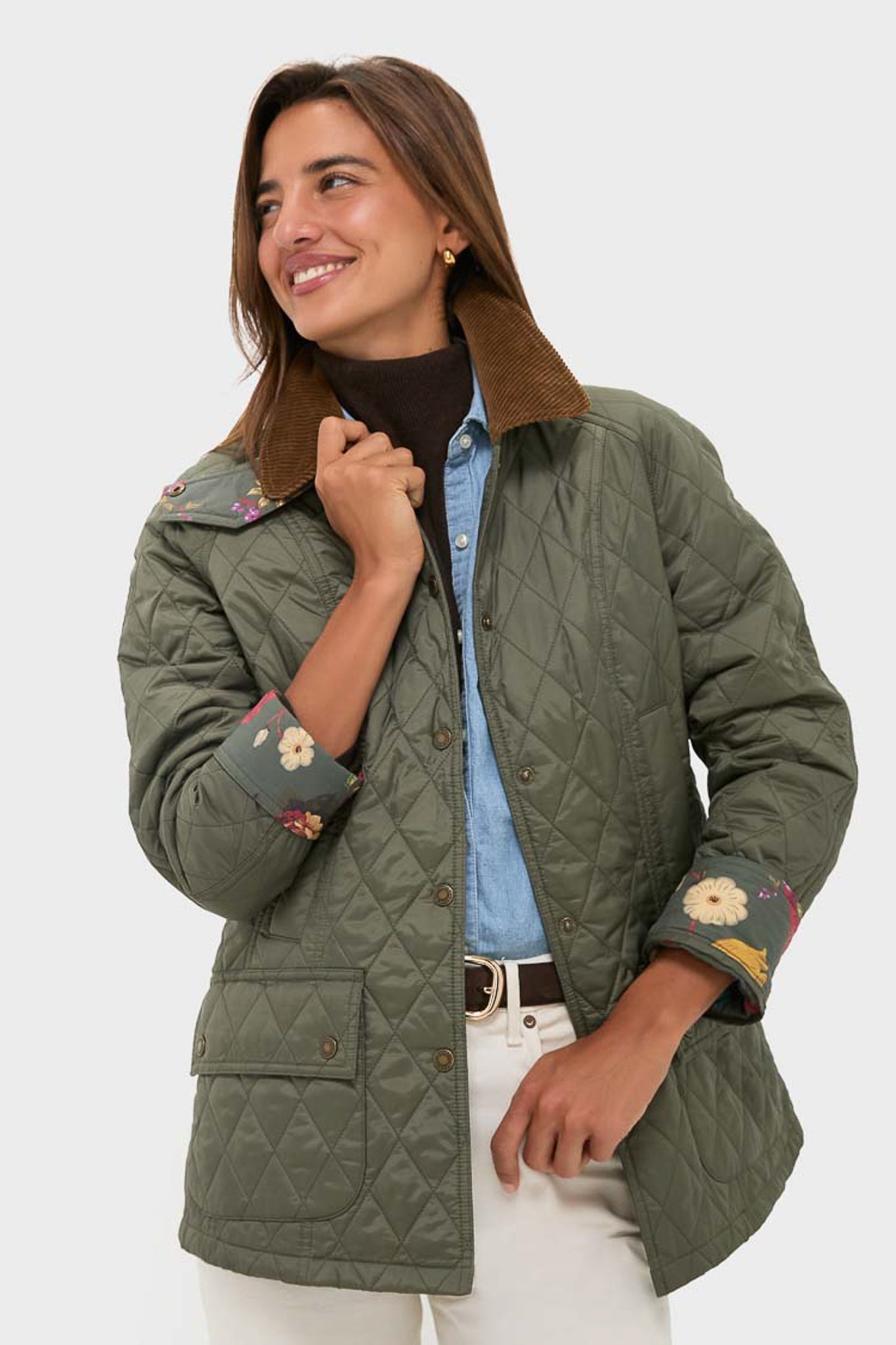 A woman smiles in a Barbour x Metrobazaarstore olive green quilted jacket with floral embroidered cuffs, paired with a blue shirt and white pants, standing against a plain background.