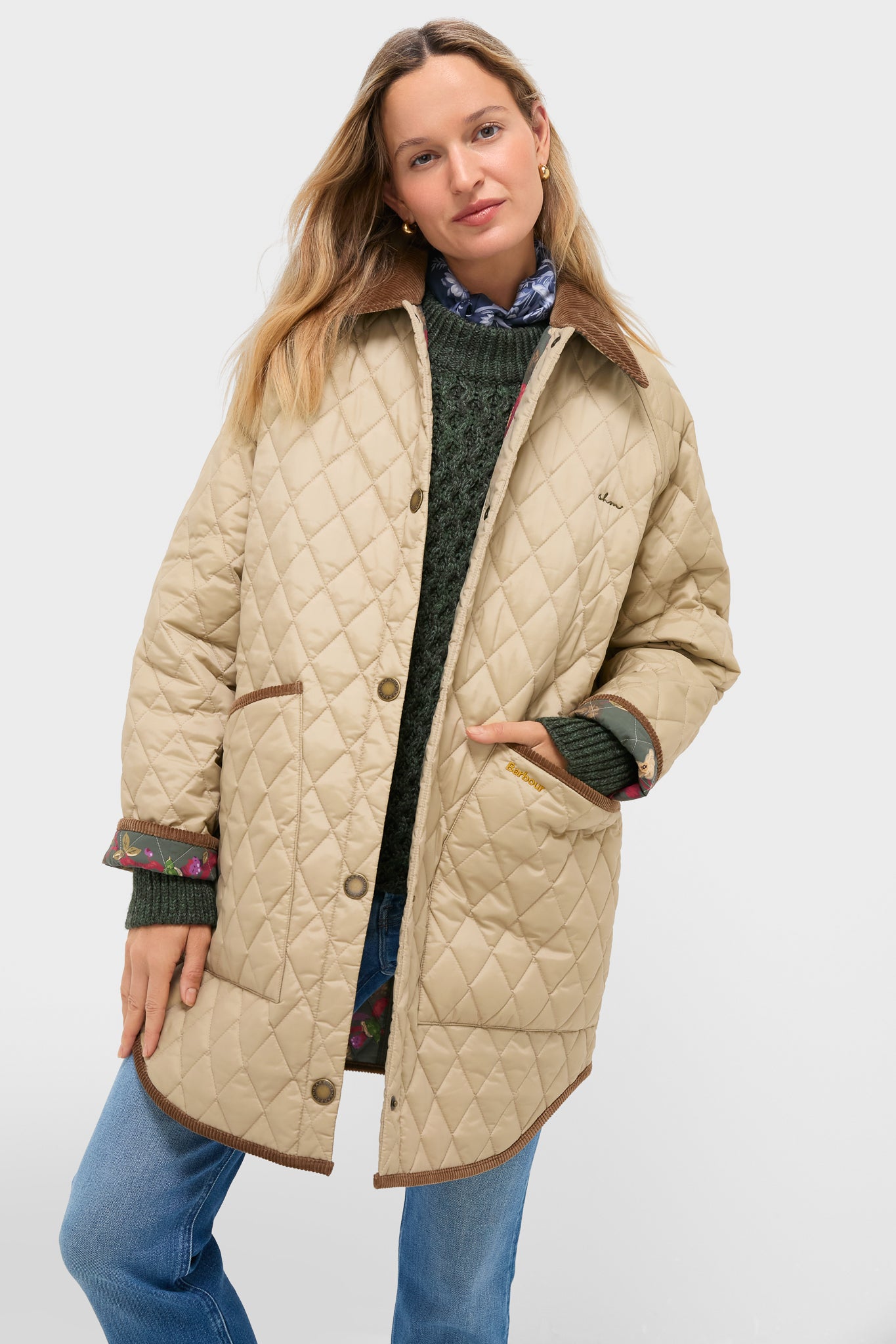 A woman wears the Barbour x Metrobazaarstore Miley quilt jacket, a beige diamond quilt style with brown trim, over a green sweater and blue jeans, standing with one hand in her pocket against a plain background.