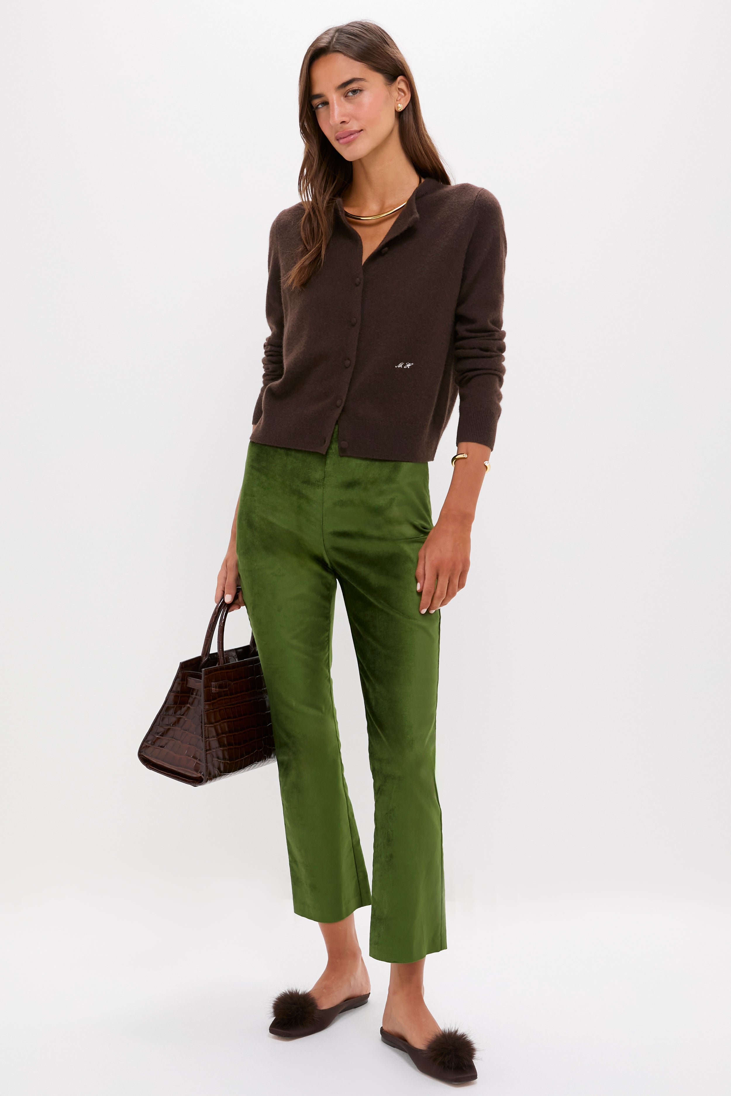 A woman dressed in Metrobazaarstore’s dark brown cardigan, high-rise green velvet kick-flare pants, and brown slippers holds a brown handbag while standing against a plain white background.