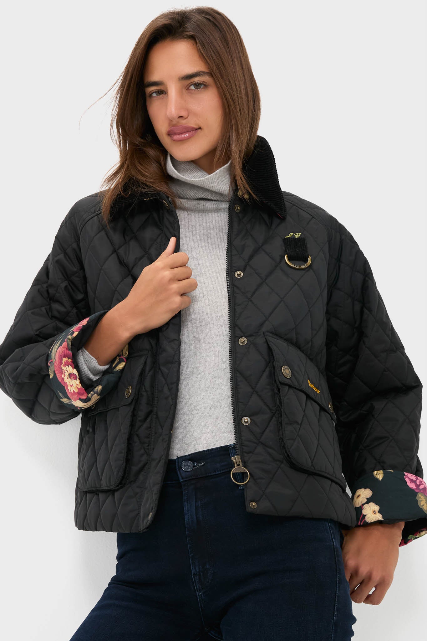 Woman in a black Barbour x Metrobazaarstore Kinsley diamond quilted jacket with floral lining, light gray turtleneck, and dark blue jeans stands against a plain white background.