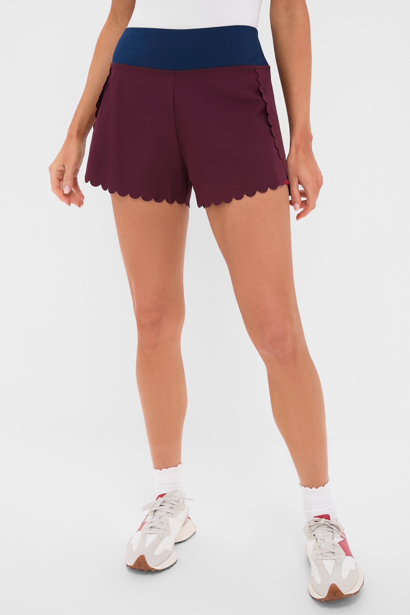 A person is wearing Tnuck Sport burgundy scalloped shorts with a navy waistband, paired with white socks and white sneakers.