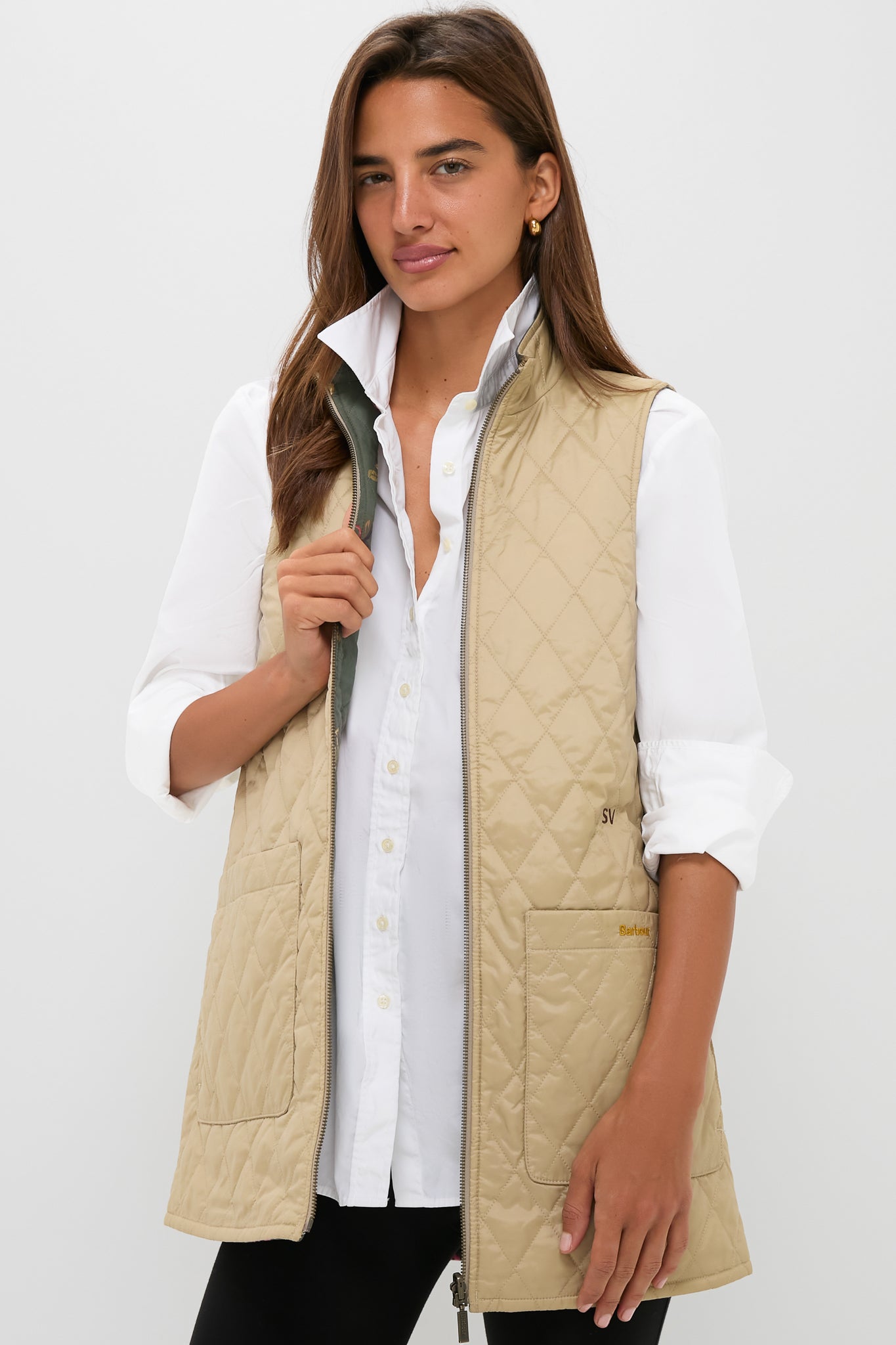 A woman stands against a white background wearing a Barbour x Metrobazaarstore beige quilted vest with a reversible paisley liner over a white button-up shirt and black pants.