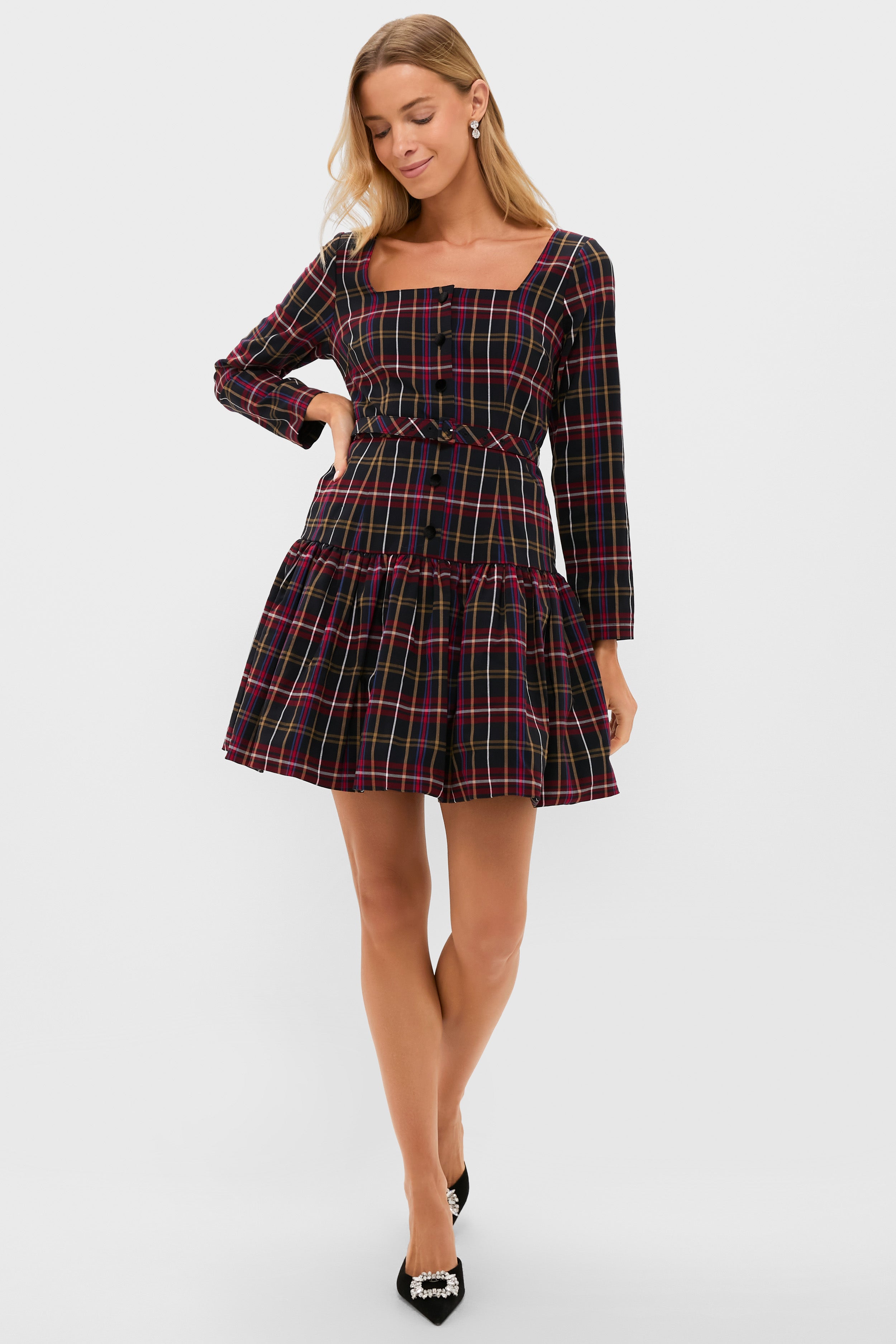 A woman wearing the Metrobazaarstore black tartan mini dress with long sleeves, a square neckline, and a tiered skirt stands with one hand on her hip, looking down—a chic choice for festive occasions.