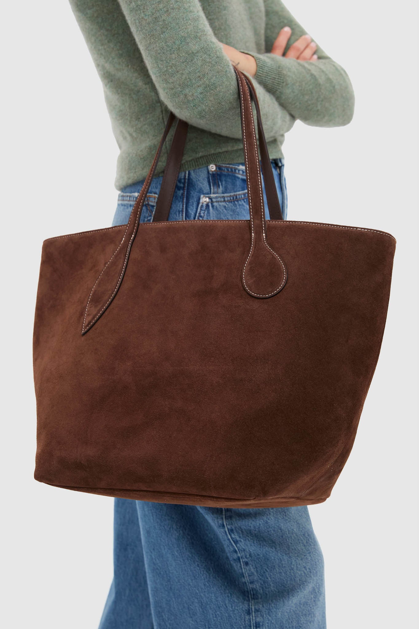 Person holding the Dark Brown Suede Large Sprout Tote, showcasing its classic, effortless design with white top-stitched handles, perfect for a fun, high-end weekend escape.