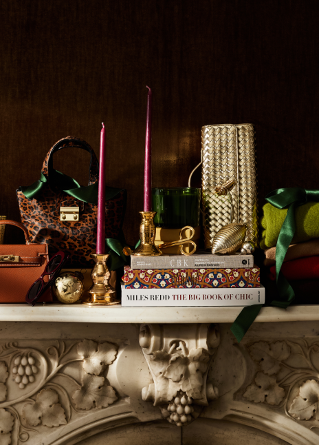 A thoughtful selection of gifts sitting on a fireplace mantel. Gifts include candles, bags, clutches, and scarves. 