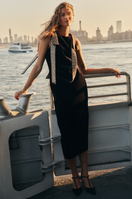 A woman stand on a boat wearing a black midi dress with embellished shoulder bows and t-strap heels.
