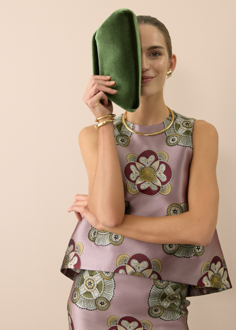 Woman holding a green velvet bag in front of half of her face in a matching lilac tank and skirt. 