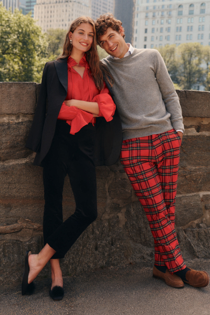 On the left, a woman stands wearing a red blouse, back pants, a black blazer, and black flats. On the right, a man wears a gray sweater over a white button down, red plaid trousers, and brown loafers.
