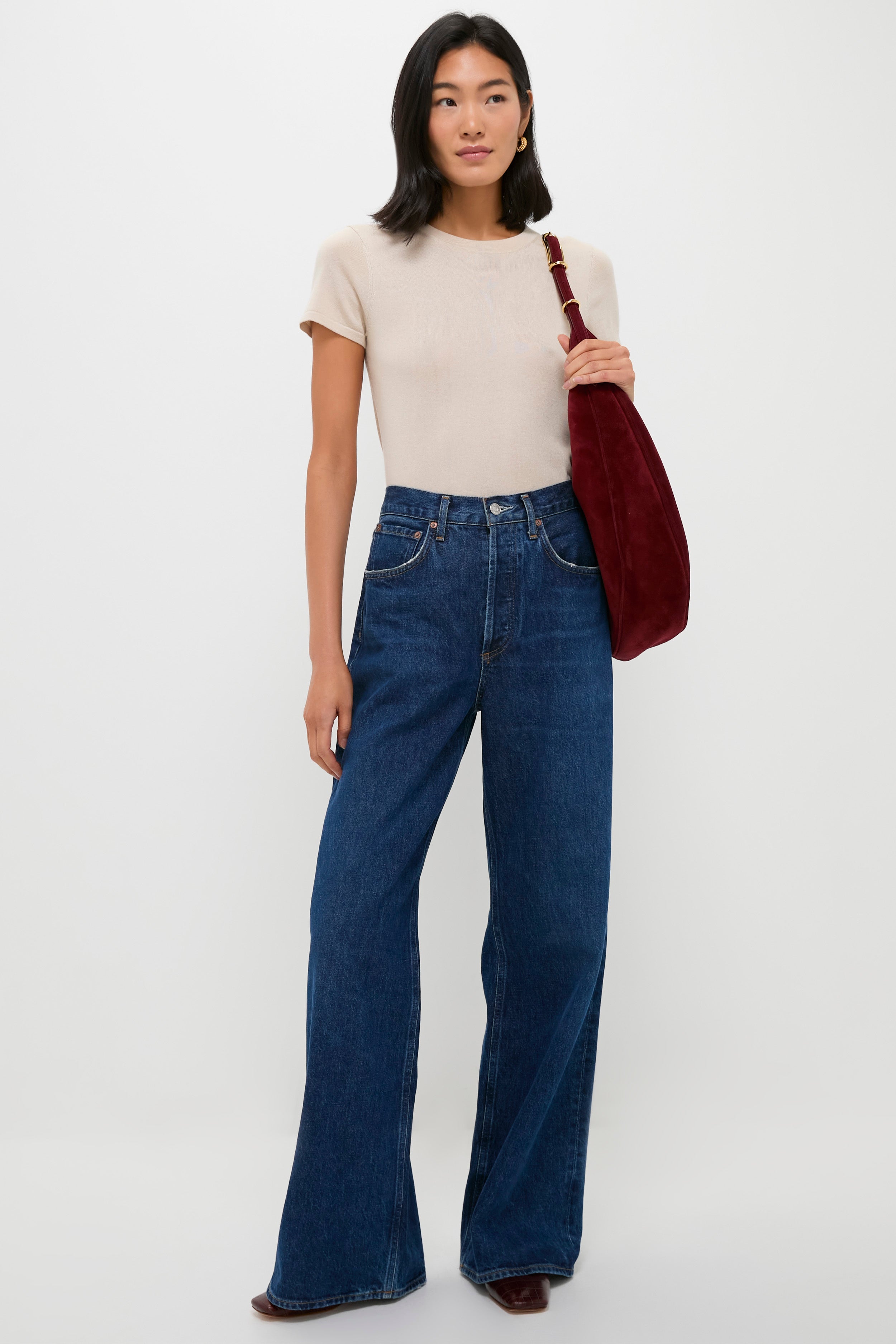 A woman stands against a plain background wearing a Metrobazaarstore beige relaxed fit crewneck t-shirt, wide-leg blue jeans, dark boots, and carrying a large dark red shoulder bag.