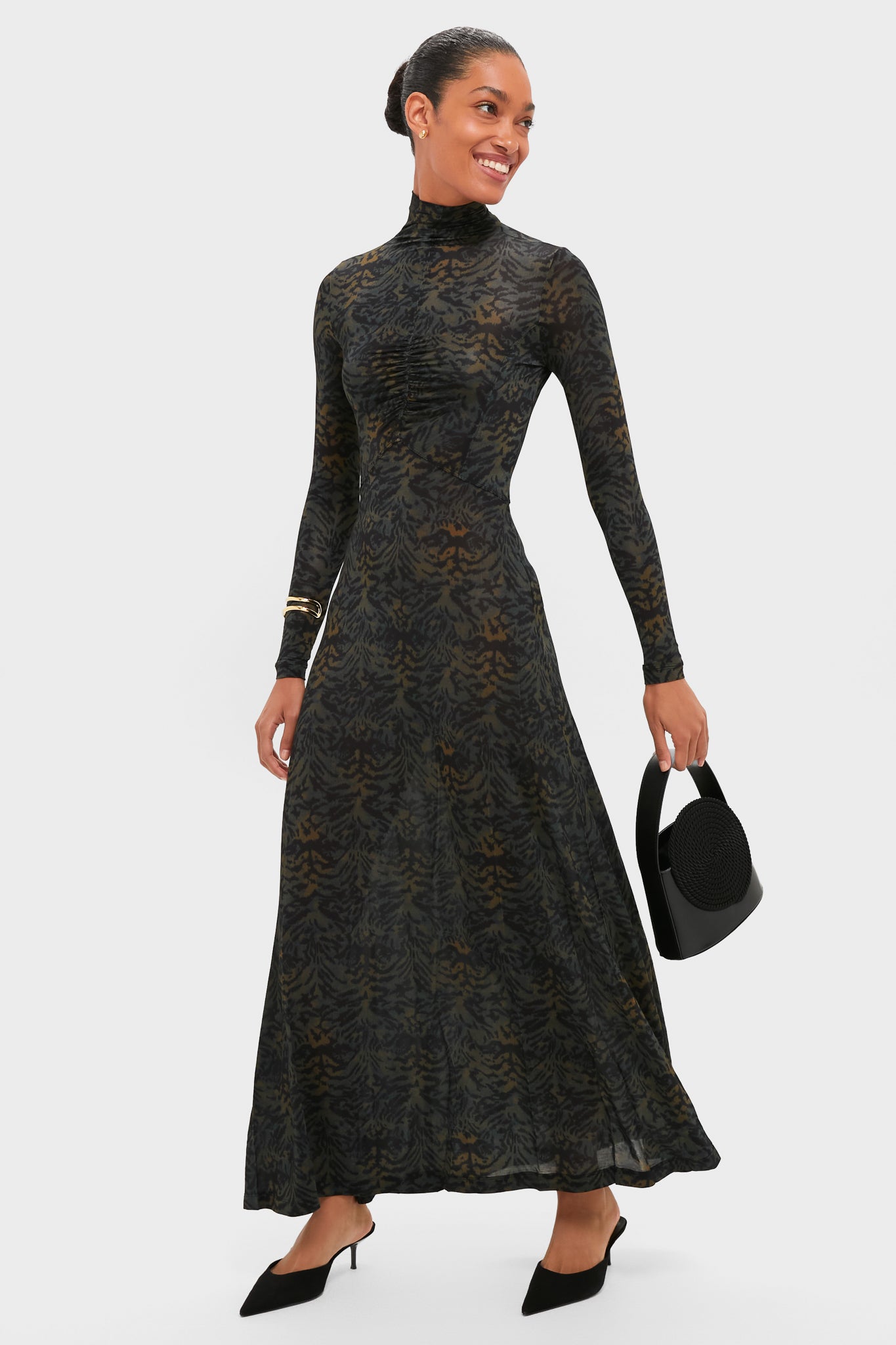 A woman in a long-sleeved, ankle-length patterned dress with black heels and a black round handbag smiles against a plain background, accessorized with Lie Studio gold-plated sterling silver Franka Earrings.