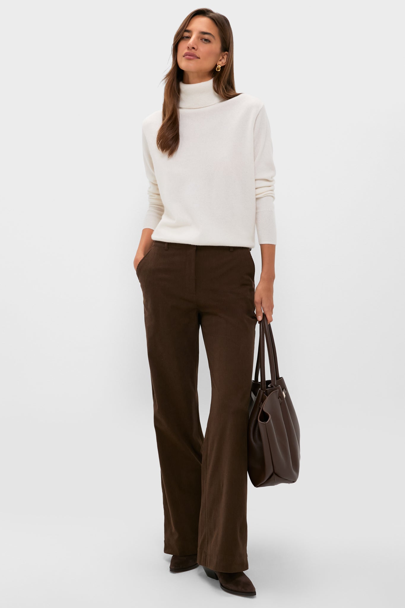 A woman stands against a plain light background wearing a Metrobazaarstore cashmere turtleneck sweater, brown wide-leg pants, dark shoes, and holding a brown tote bag.