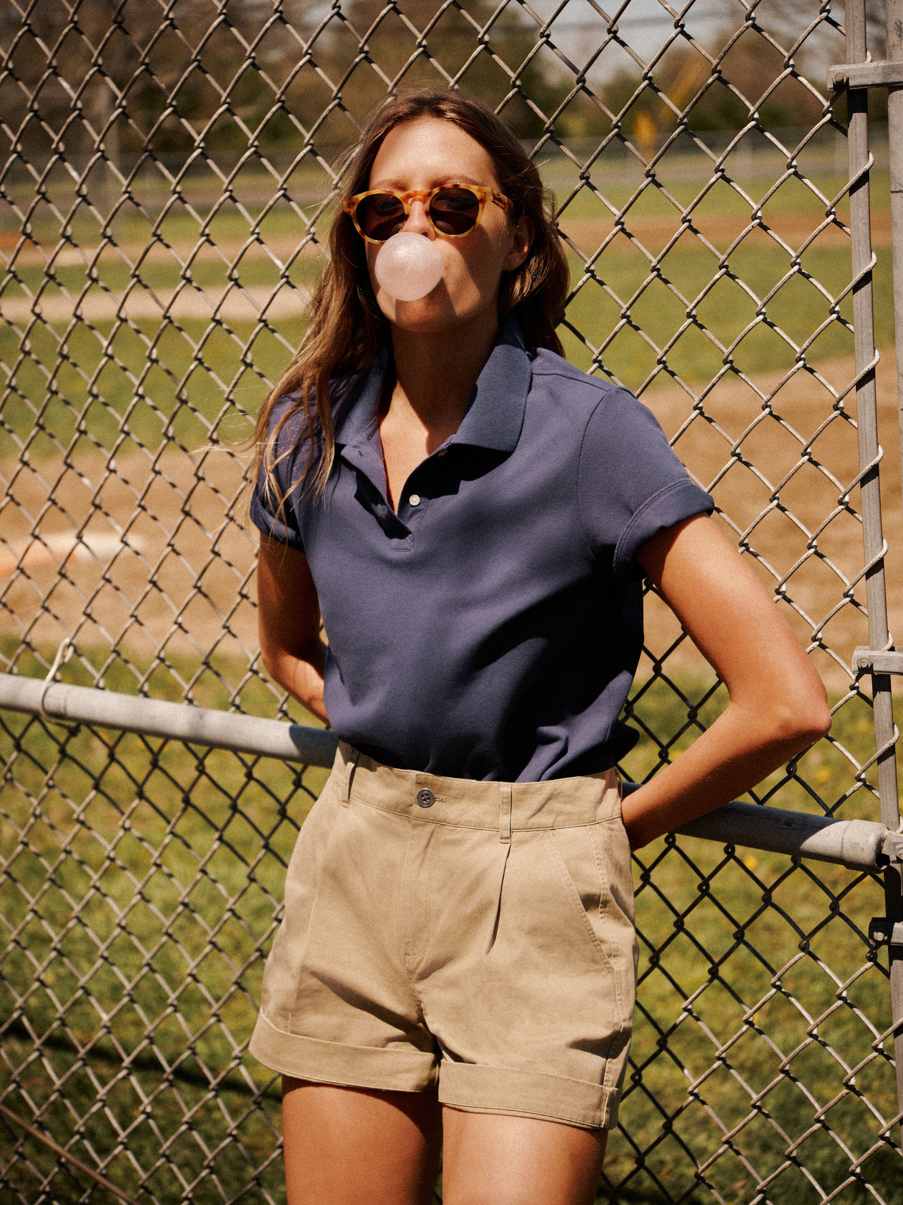 A person in Metrobazaarstore sunglasses, a blue Holmes Polo in textured pique fabric, and khaki shorts stands by a chain-link fence, blowing a gum bubble.