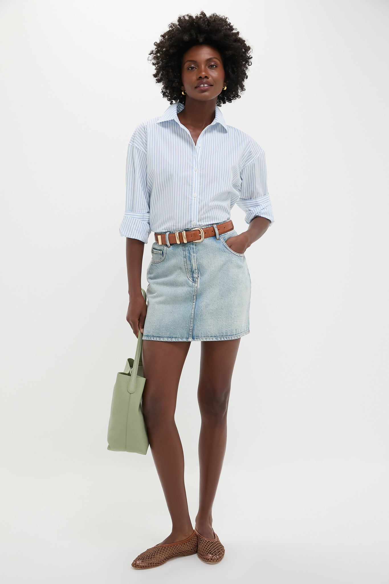 A woman stands against a white background wearing a light blue striped Metrobazaarstore Wexler Button Down with drop shoulders, a denim mini skirt, brown belt, woven brown flats, and carrying a light green handbag.