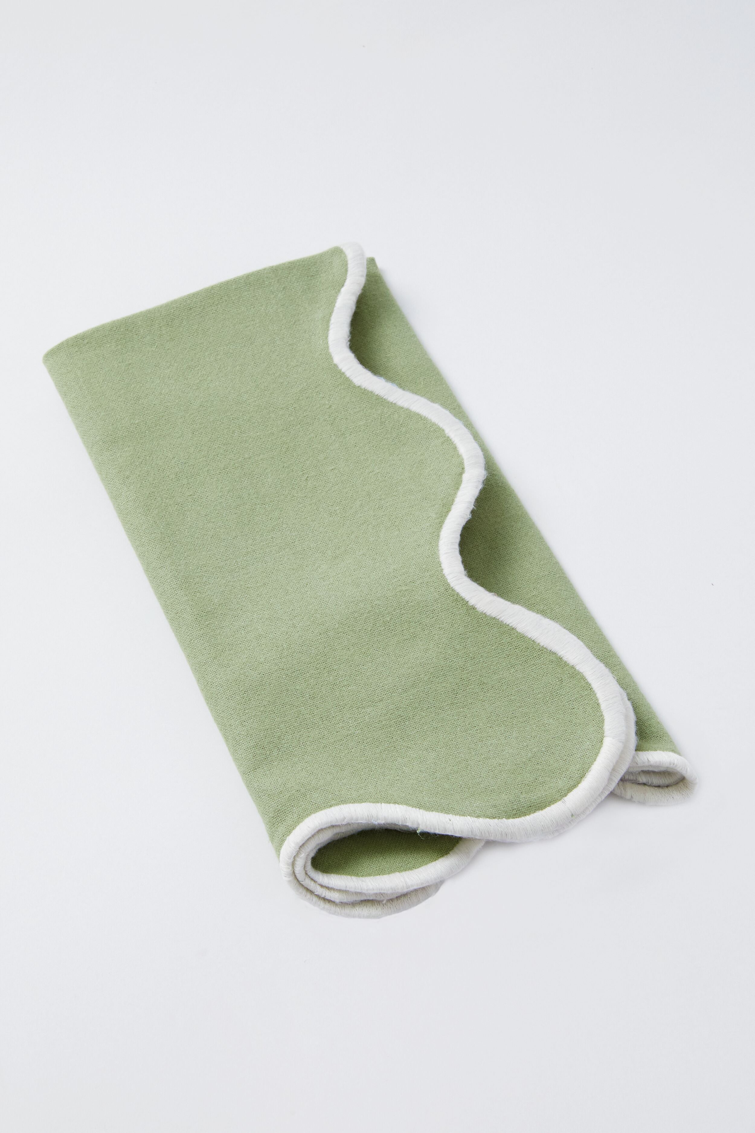A folded light green Metrobazaarstore Home blanket with white trim, displayed on a white surface, pairs beautifully with scalloped napkins for an elegant touch.