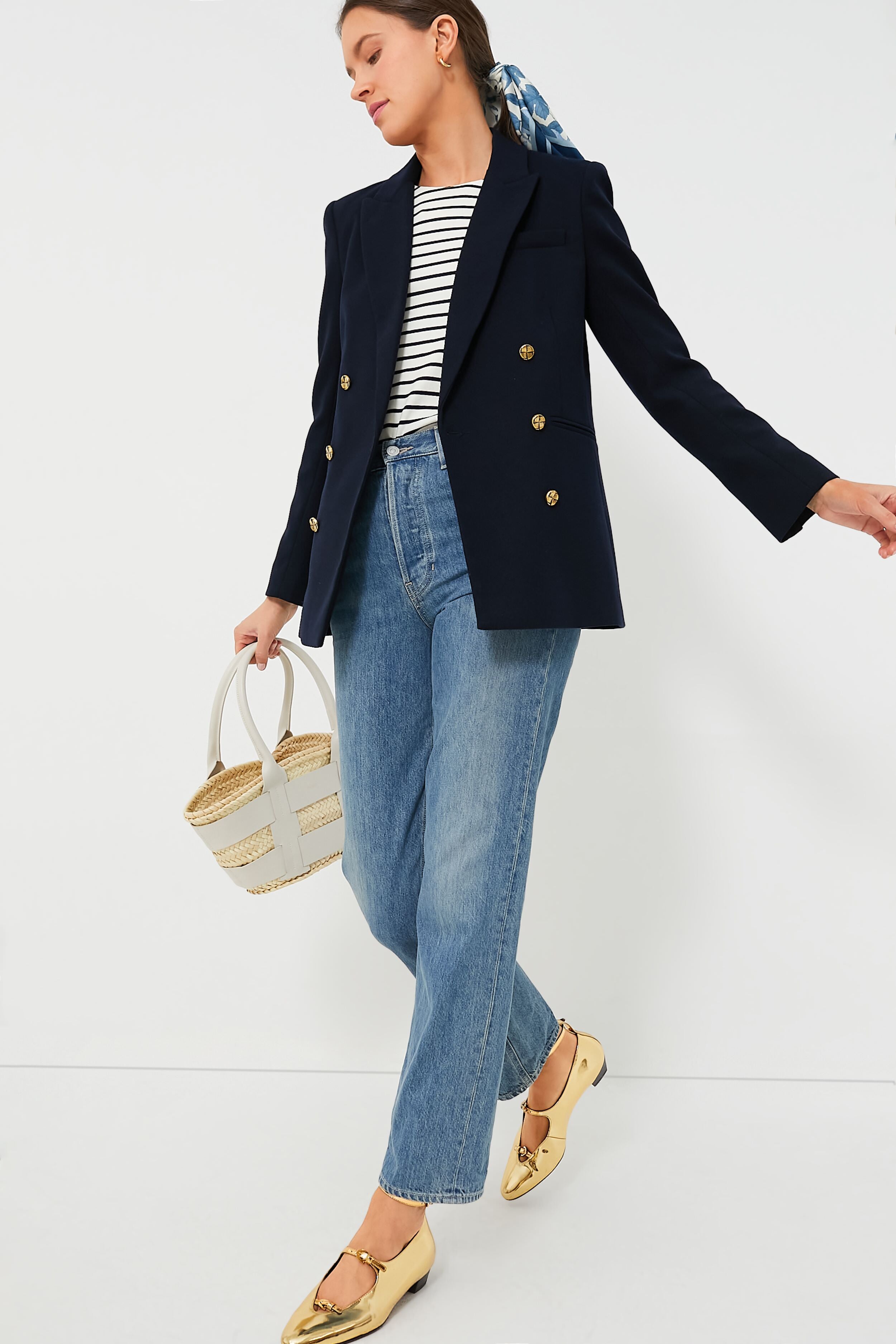 A person wears a Metrobazaarstore Navy Diana Blazer with gold buttons, a striped top, blue jeans, gold flats, and carries a woven handbag.